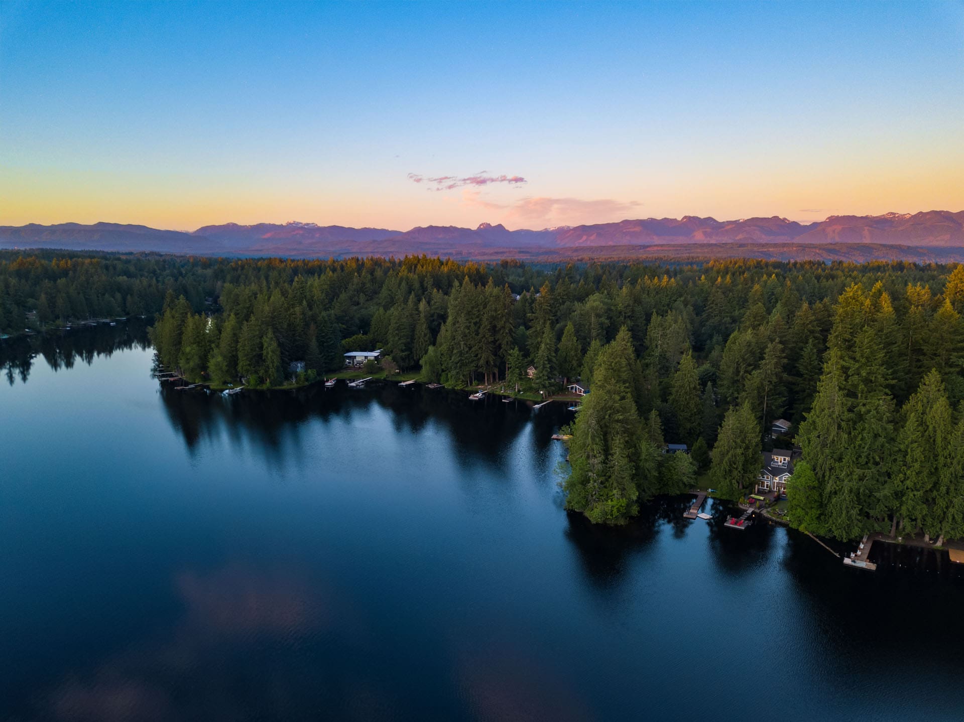 A serene lake is bordered by a dense forest of evergreen trees. The setting sun casts a warm glow across the sky, creating a gradient from orange to blue. Distant mountain ranges are visible on the horizon, enhancing the tranquil and picturesque landscape.