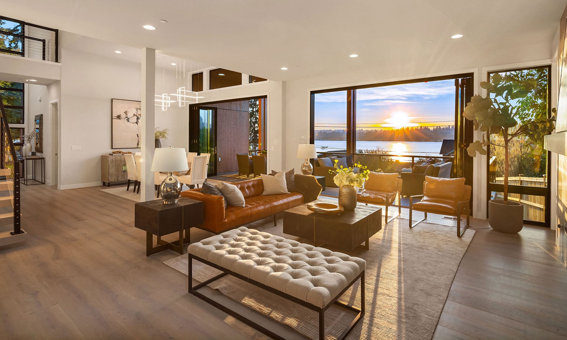 A modern living room with a sunset view over a balcony. The room features a large tan leather sofa, a cushioned bench, wooden coffee tables, and decorative plants. Floor-to-ceiling windows and sliding doors allow natural light to illuminate the sleek interior design.