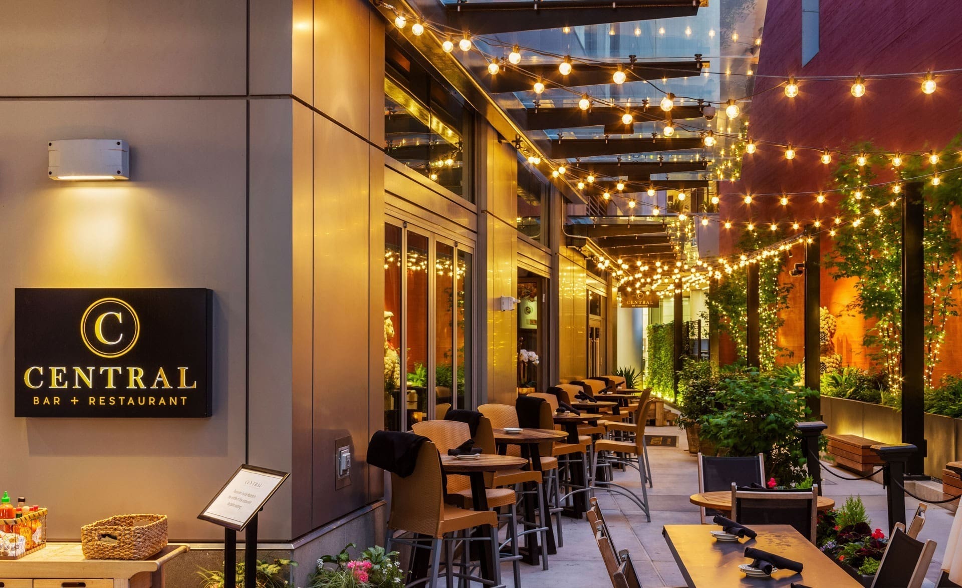 An outdoor seating area of a restaurant with yellow string lights hanging overhead, creating a cozy ambiance. Bar stools, high tables, and lush green plants line the narrow patio. A sign reads "Central Bar + Restaurant" on the left wall.