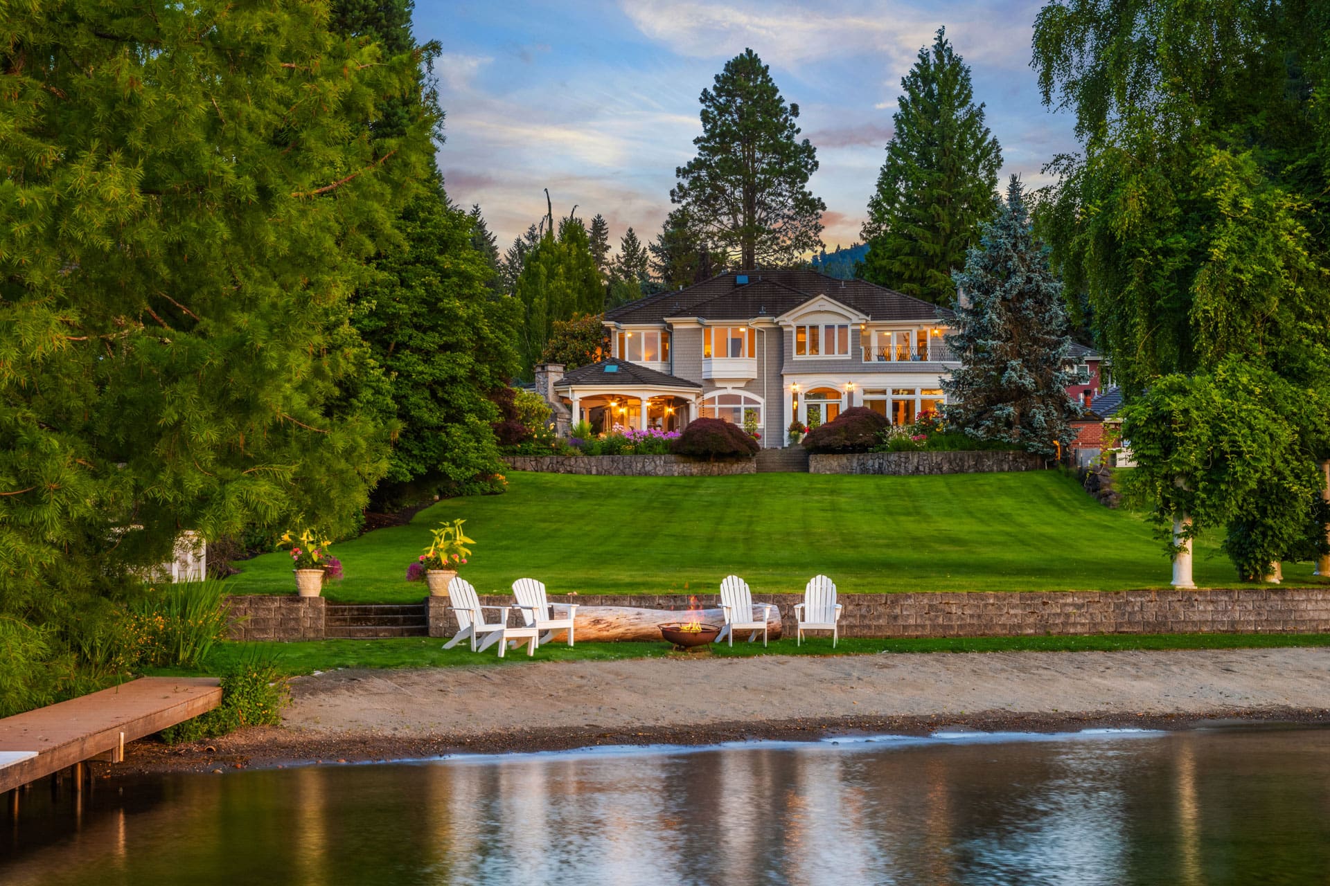 A large, elegant two-story house with multiple windows is surrounded by lush gardens and tall trees. At the edge of a manicured lawn that extends to a calm lakeshore, white Adirondack chairs encircle a fire pit near a small dock. The scene is serene and picturesque at dusk.