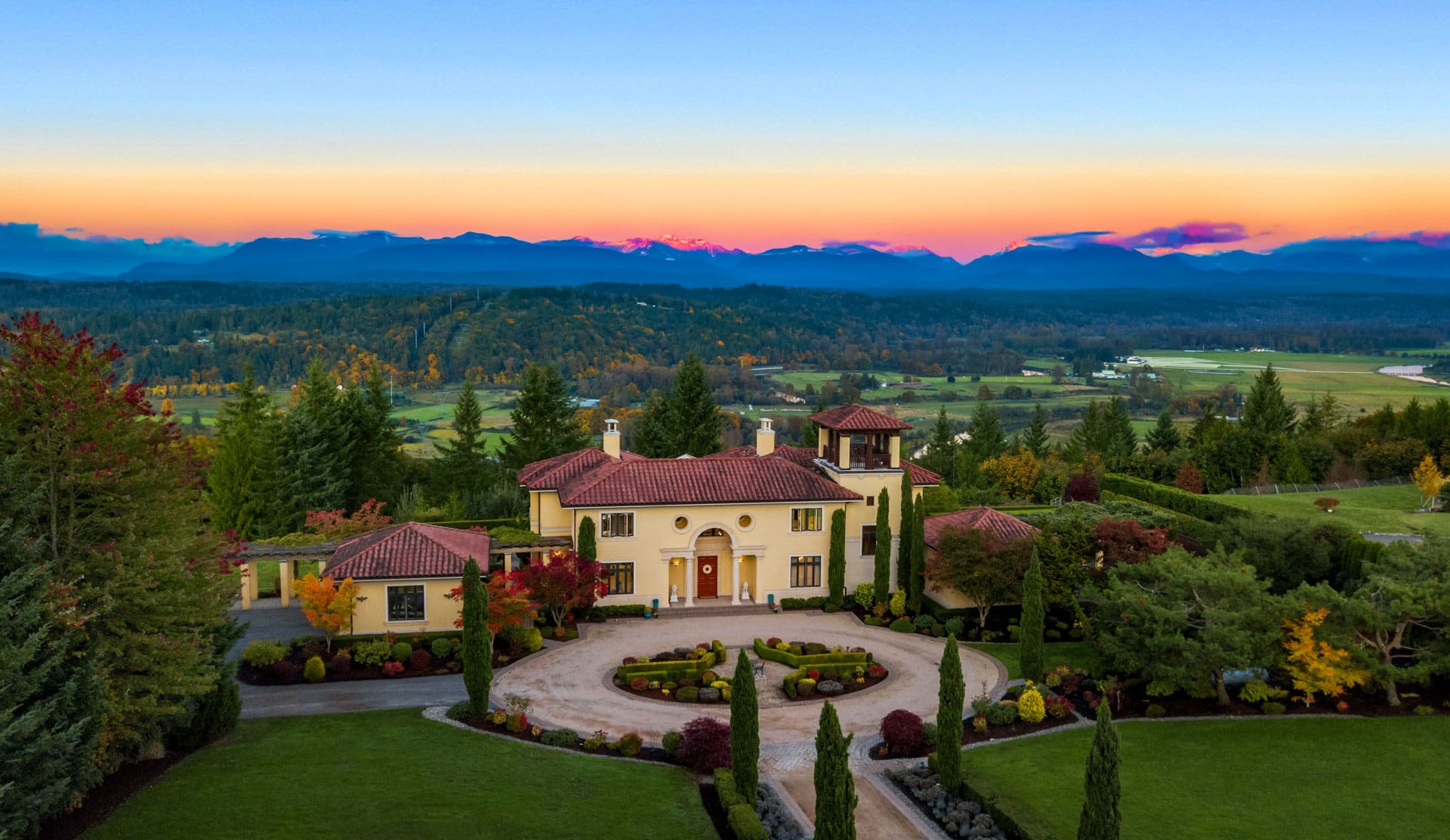 An aerial view of a large, luxurious Mediterranean-style villa with red-tiled roofs and cream-colored walls, surrounded by manicured gardens and lush greenery. A scenic countryside and forest extend into the distance under a sky transitioning from evening to night.