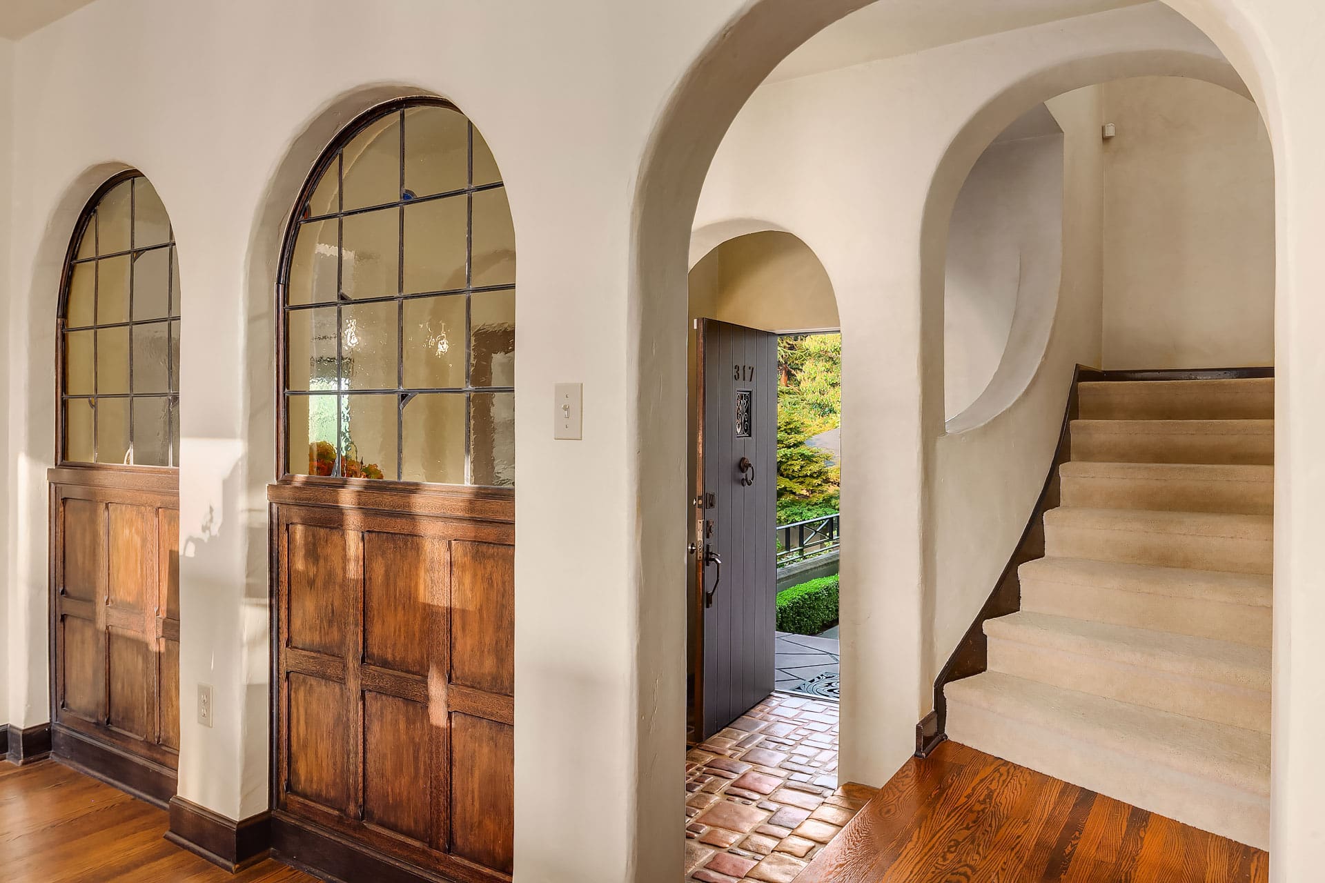 A warmly lit hallway with smooth white walls features three large arches. An open door reveals the exterior view. A staircase on the right leads upstairs, and wooden paneled windows allow light to stream in, complementing the wooden flooring.