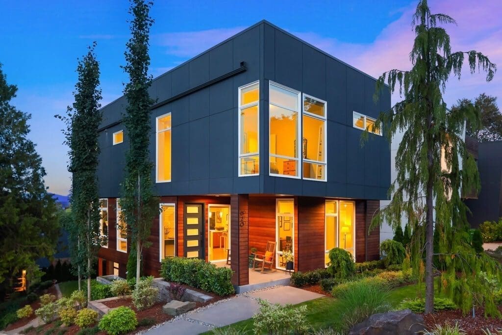 A modern two-story house with large windows illuminated from inside. The exterior features a combination of dark panels and wood accents. The house is surrounded by neatly landscaped greenery and tall, slender trees, under a clear evening sky.