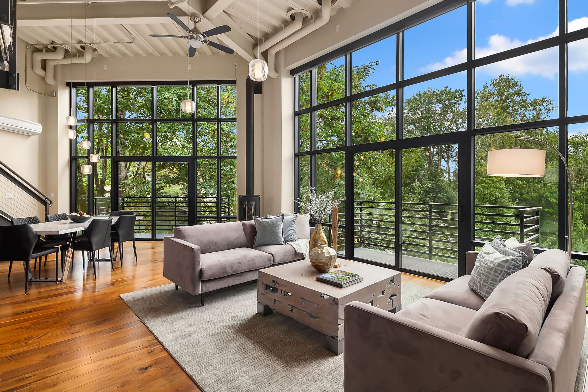 A modern living room with large floor-to-ceiling windows showcasing a lush green view. The room features two gray sofas, a wooden coffee table, and pendant lights. The dining area with a black table and chairs is visible to the left, along with a staircase.