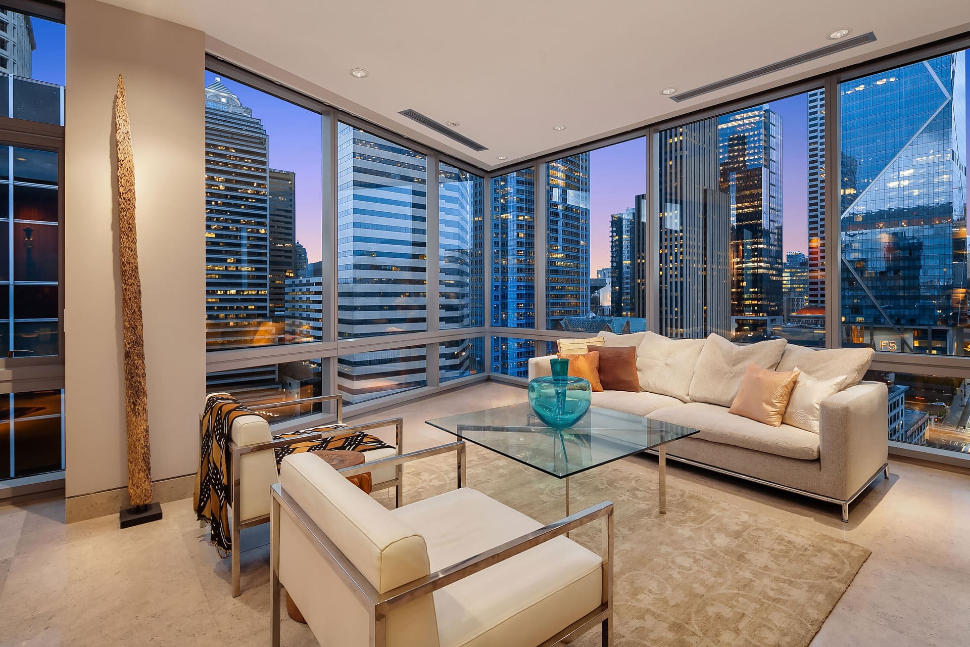 A modern, luxurious living room with floor-to-ceiling windows showcasing a cityscape view. The room features a white sofa, a glass coffee table with a blue decorative vase, a white armchair, and a minimalist aesthetic accented by a tall, slender sculpture.