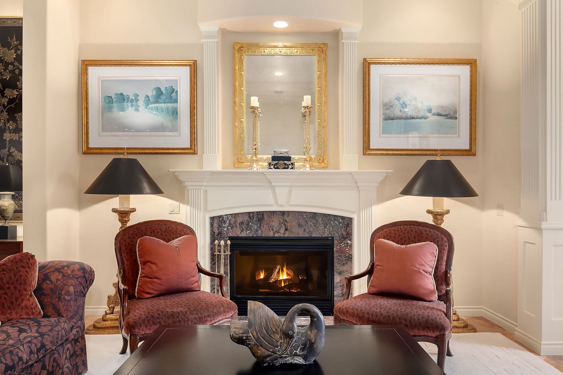 A cozy, elegant living room features a lit fireplace with a wooden mantle, flanked by two framed paintings and gold candleholders. Two ornate red armchairs with matching cushions face a black coffee table adorned with a decorative swan sculpture.