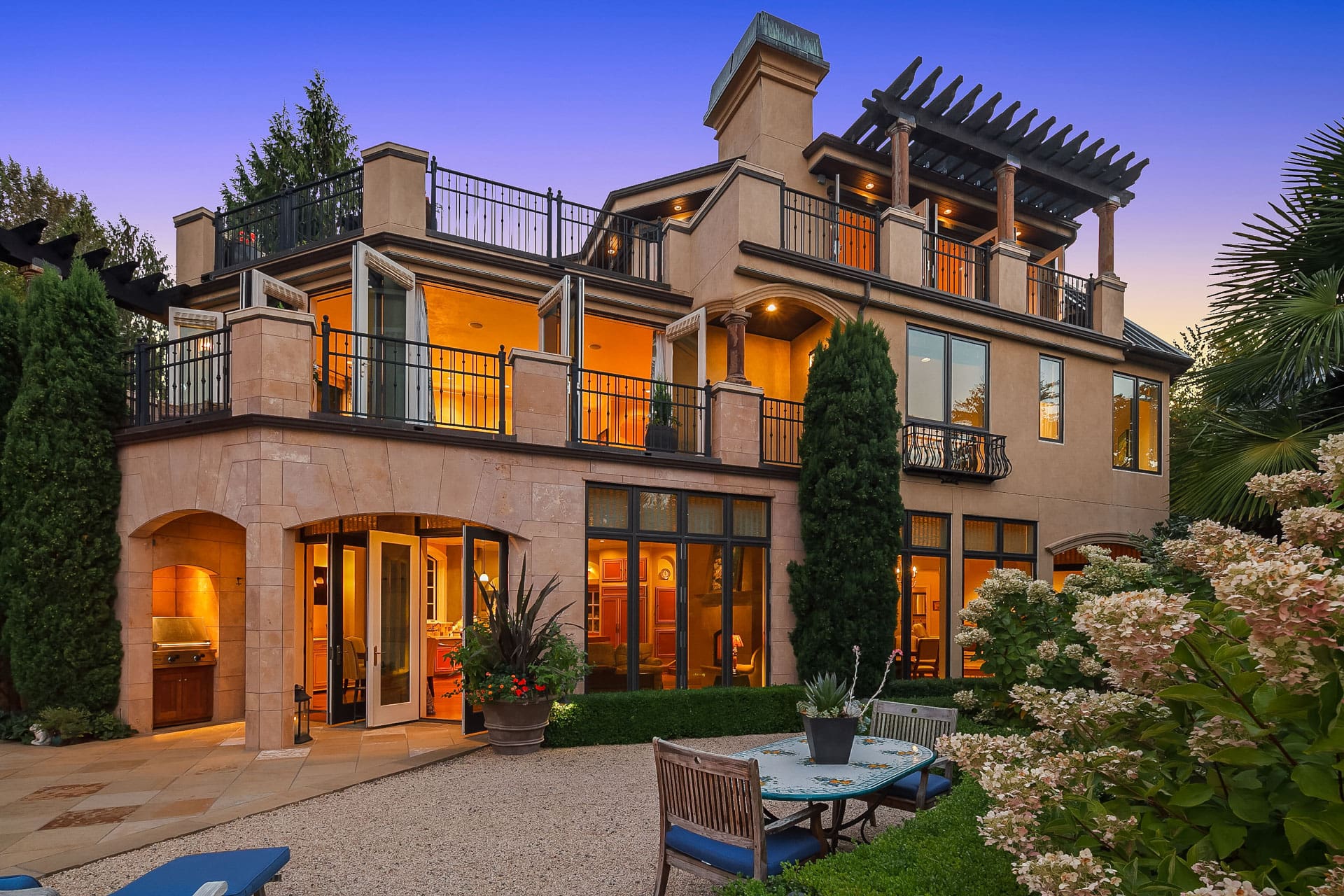 A luxurious two-story house with large windows and multiple balconies, adorned with warm exterior lighting. The patio area features outdoor seating, lush greenery, and a pergola on the rooftop, set against a twilight sky.