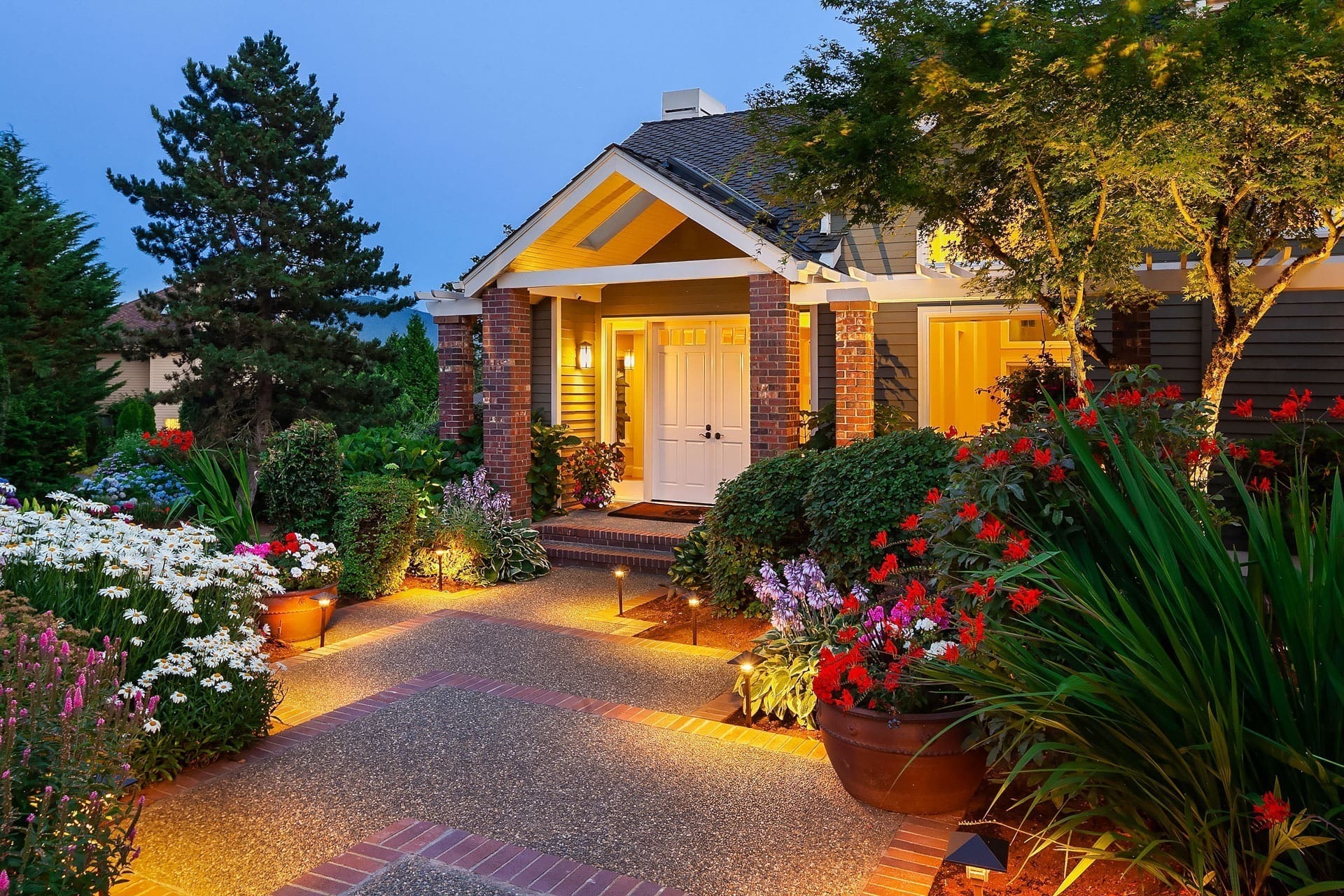 A well-lit pathway leads to a charming house with a gabled roof and brick accents at dusk. The entrance is adorned with potted flowers and colorful garden beds, creating a vibrant and welcoming atmosphere. Trees and shrubs surround the house, blending beautifully with the setting.