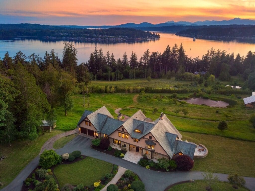 Aerial view of a large, luxurious house surrounded by lush greenery, with a scenic lake and forested hills in the background at sunset. The house features a landscaped garden and a winding driveway. The sky is painted with warm hues of orange and pink.