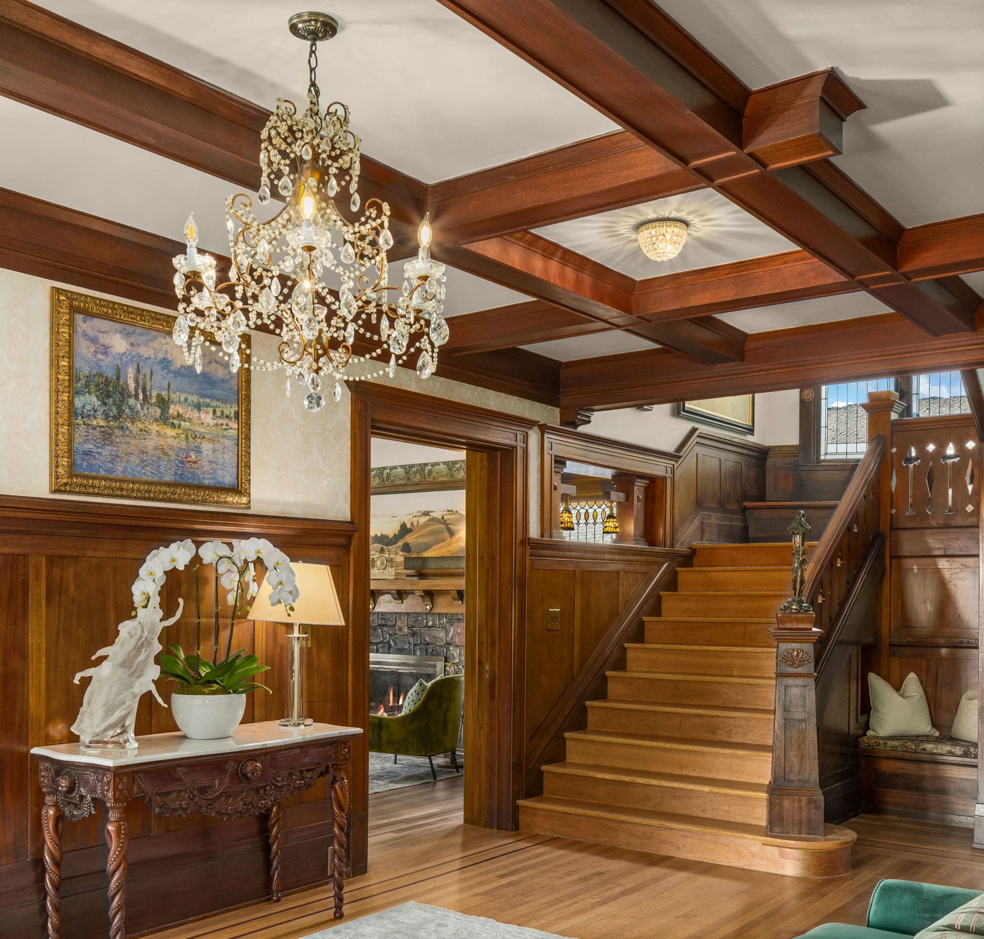 A luxurious foyer features a wooden staircase, a crystal chandelier, and an ornate table holding a large white orchid in a pot. The room has rich wood paneling, a painting on the wall, and a glimpse of a cozy room with a fireplace in the background.