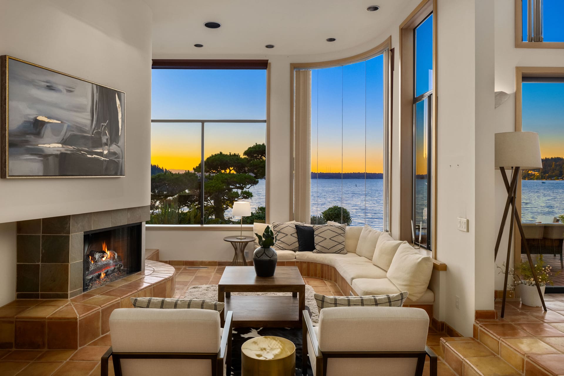 A modern living room with large windows showcasing a waterfront sunset view. The room features a sectional sofa, two armchairs, a coffee table, potted plants, and a fireplace. A floor lamp is positioned next to the sofa, and a piece of abstract art hangs above the fireplace.