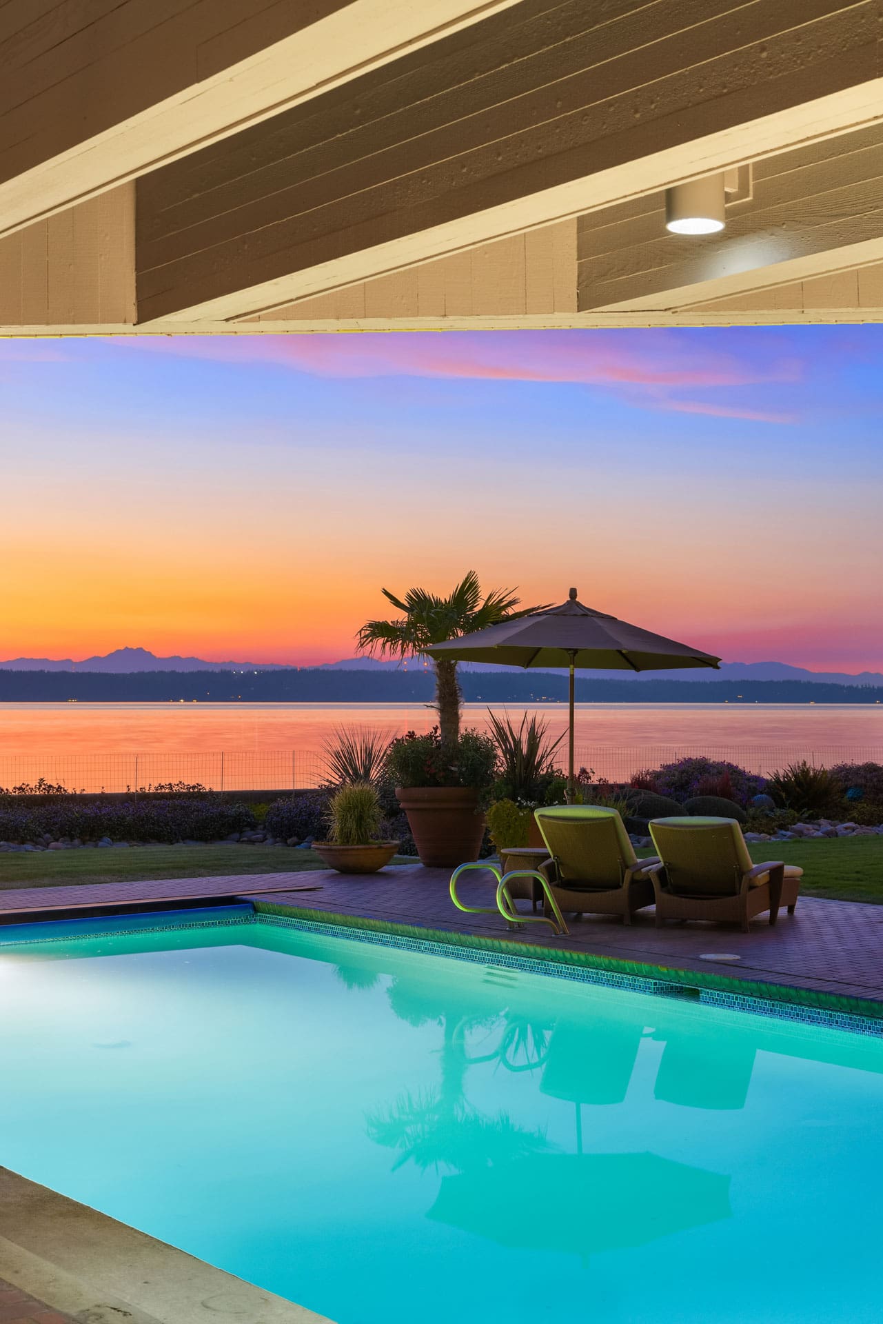 The image shows a serene backyard scene featuring a swimming pool with two lounge chairs and an umbrella next to it. Beyond the pool, there's a picturesque view of a calm lake and mountains under a vibrant sunset sky with hues of orange, pink, and purple.