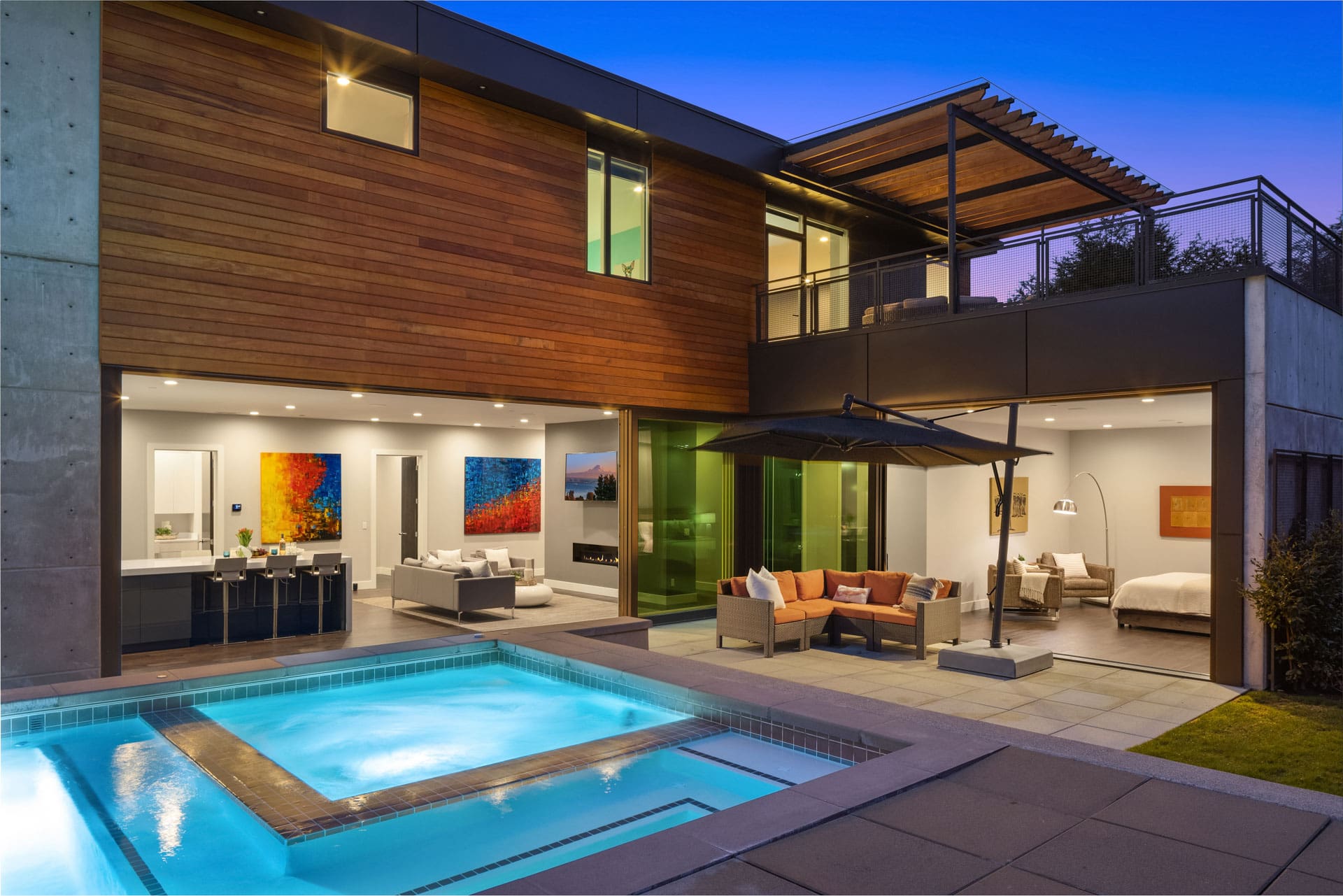 Modern two-story house at dusk with a pool in view. The ground floor features an open living area with large windows, contemporary furniture, colorful artwork, and a sleek kitchen. The patio has seating, an umbrella, and softly glowing pool lights enhance the serene setting.