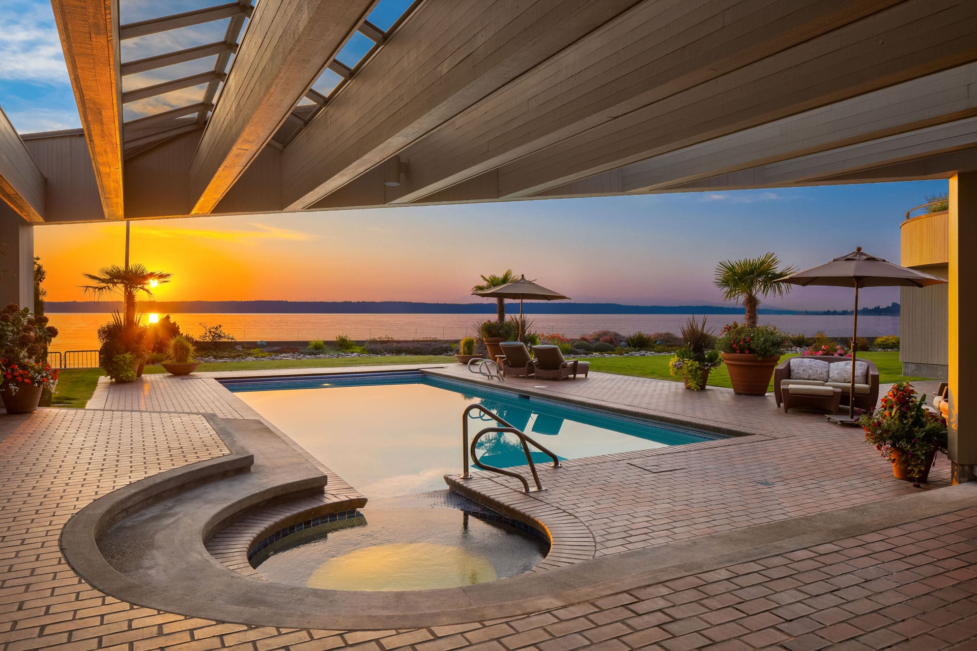 Indoor swimming pool and jacuzzi with an open ceiling and a view of a sunset over a calm body of water. Lounge chairs, umbrellas, and lush plants surround the pool, creating a serene and luxurious atmosphere.