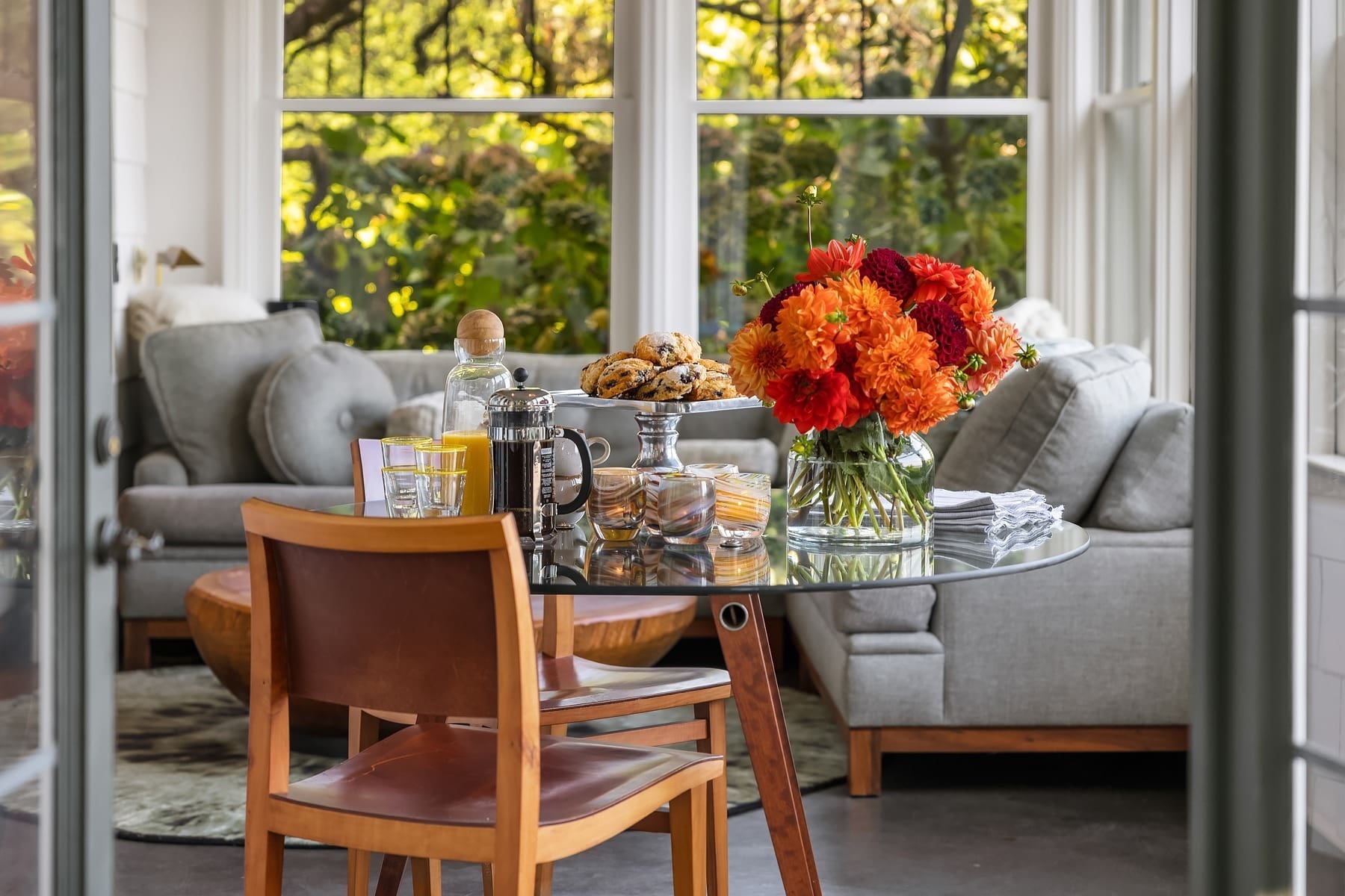 A cozy sunlit room features a round glass table set with a French press, juice, glasses, and a vase of orange flowers. A plate of cookies sits on a stand. Behind the table, there’s a light grey couch adorned with cushions, all surrounded by large windows with lush greenery outside.