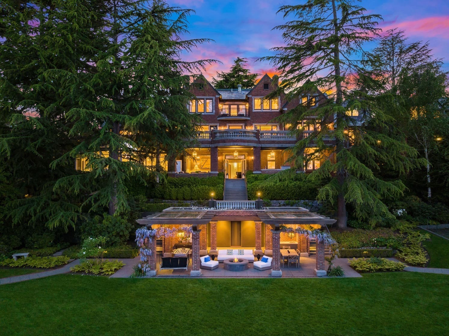 A luxurious, multi-story brick mansion with numerous large windows is illuminated at dusk. Surrounded by lush greenery, it features a spacious, well-lit patio area with seating, manicured lawns, and tall trees in the background against a vibrant sunset sky.