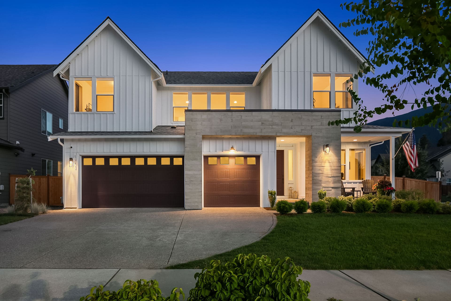 A modern two-story house at dusk with white siding, large windows, and two dark garage doors. The home features a well-lit exterior, a manicured front lawn, and a small porch area with outdoor furniture. An American flag is displayed near the entrance.