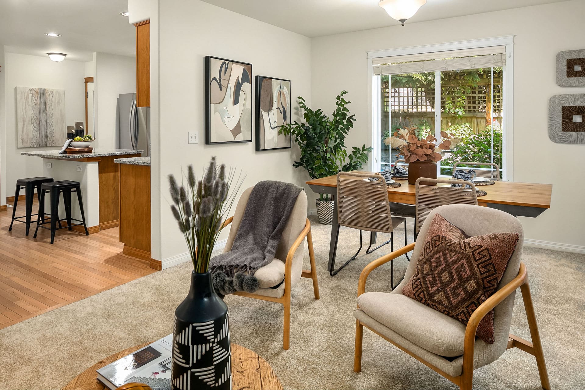 A cozy living and dining area with a neutral color scheme. The room features two cushioned chairs with patterned pillows, a wooden table with decorative items, framed art on the walls, and a large window revealing a lush garden outside. A kitchen is visible in the background.