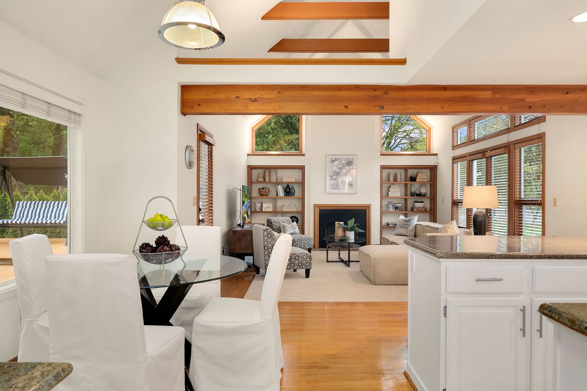 A bright and airy living area featuring a dining table with white-covered chairs, a kitchen island with white cabinetry, and a cozy seating area with a fireplace. The space has large windows with wooden frames and a high ceiling with exposed wooden beams.