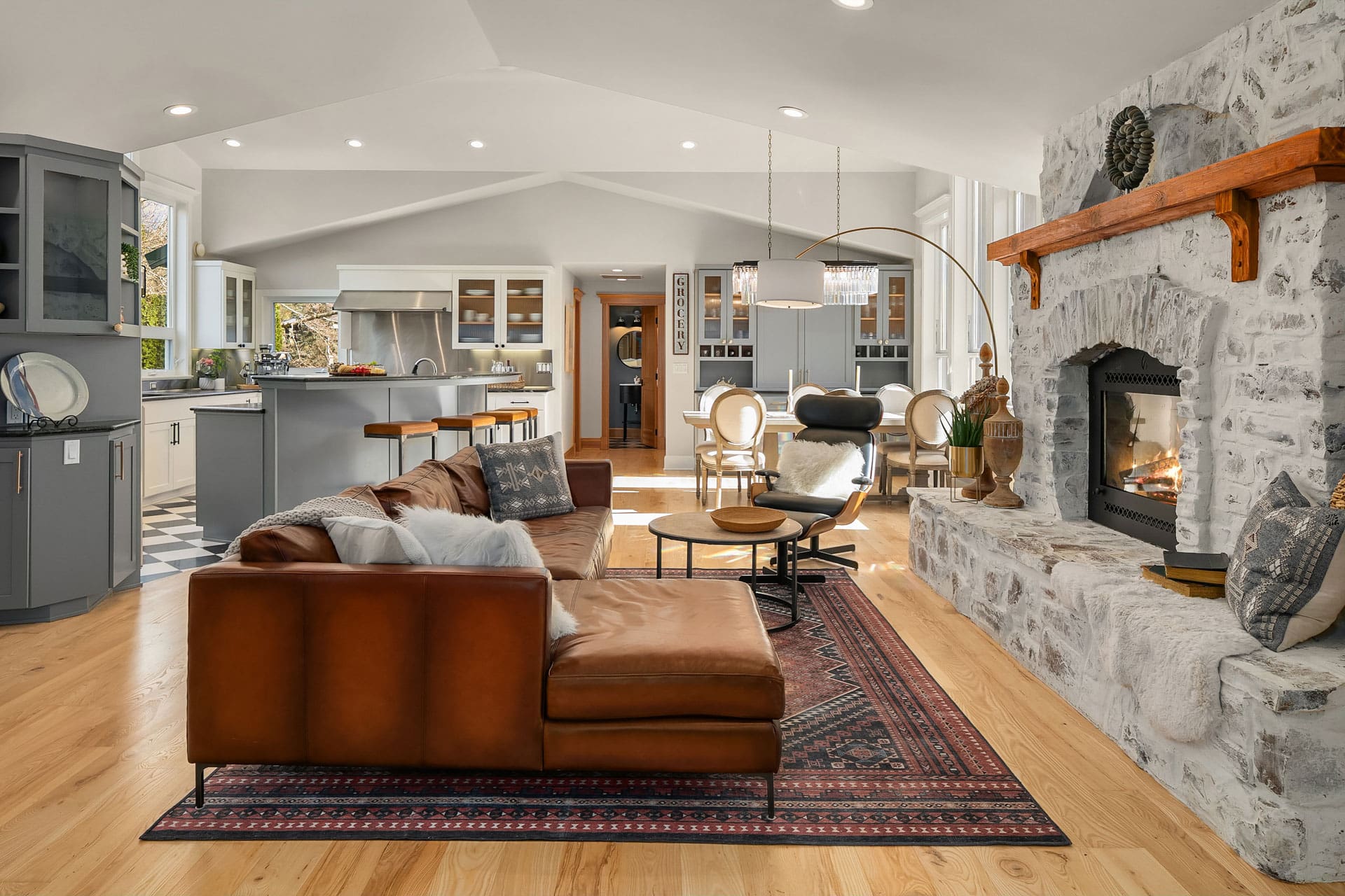 Spacious modern living room with an open floor plan to the kitchen. The room features a brown leather sofa, stone fireplace, wooden flooring, and grey cabinetry. A large patterned rug, various seating options, and abundant natural light enhance the cozy atmosphere.