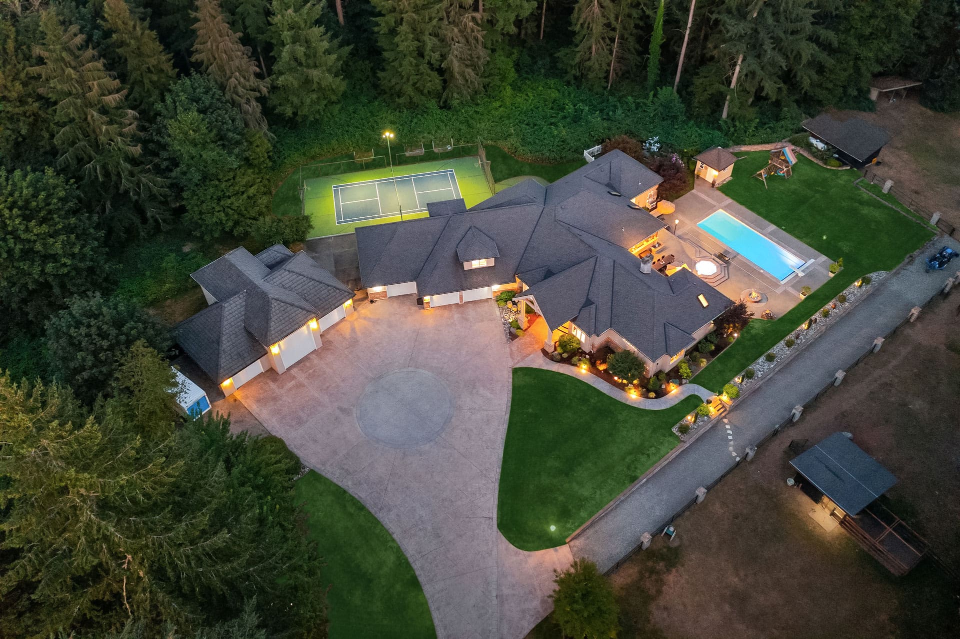 Aerial view of a large property featuring a modern house with a circular driveway, well-maintained lawn, and various outdoor amenities. There's a tennis court, swimming pool, hot tub, and multiple seating areas surrounded by lush greenery and trees.