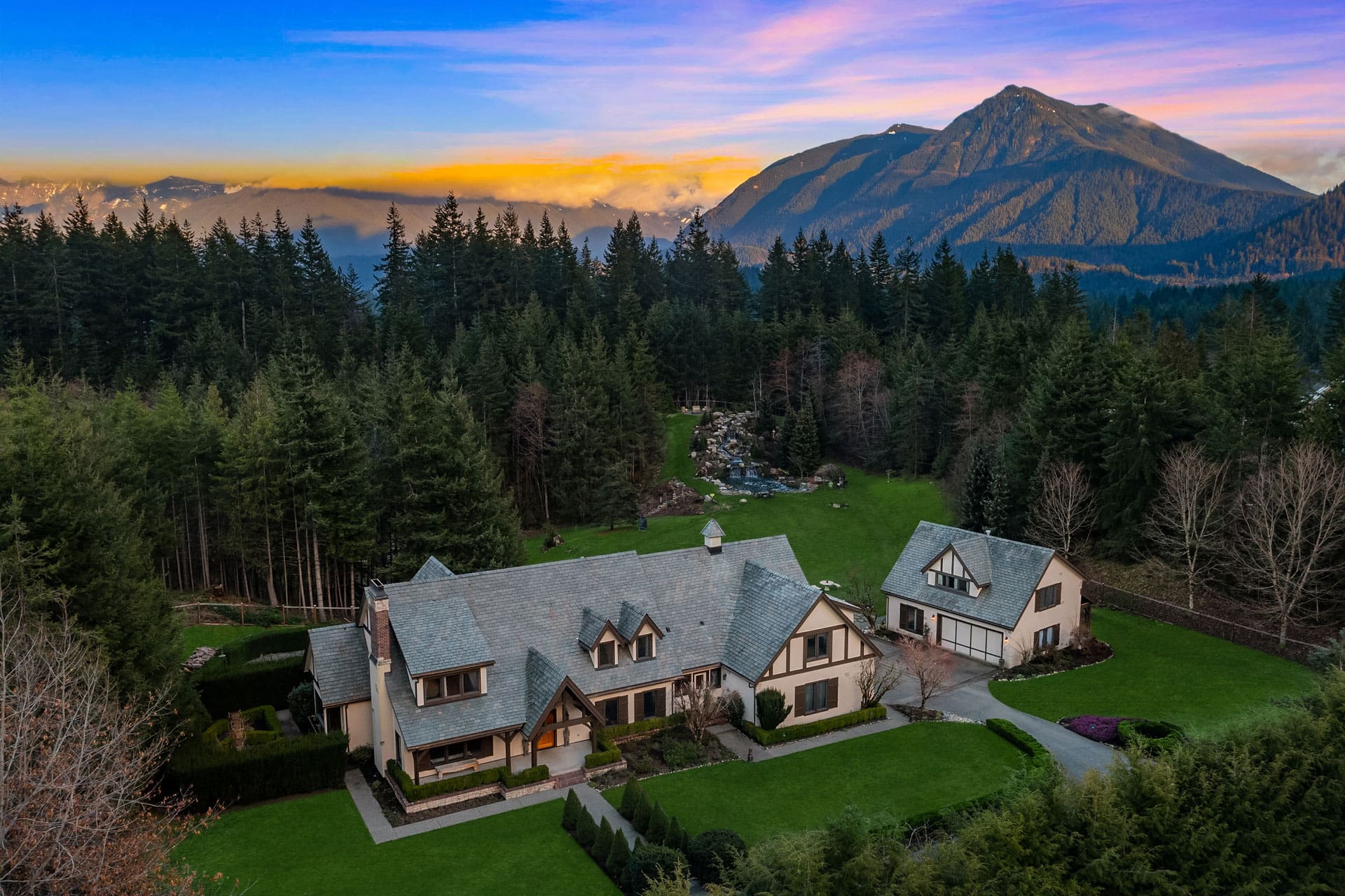 Aerial view of a picturesque property featuring a large house with multiple gables and a smaller adjacent building, both with steep roofs. The property is surrounded by lush green lawns, dense forests, and a backdrop of towering mountains under a vibrant sunset sky.