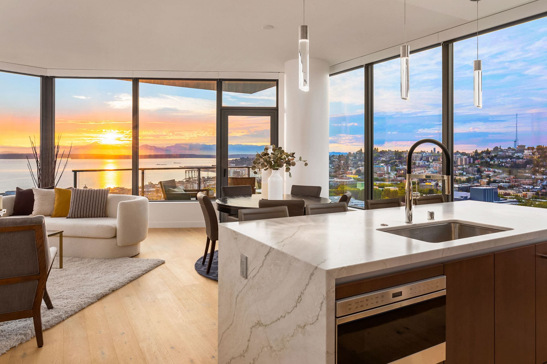 Modern open-plan living area with large windows providing a spectacular view of a sunset over water and a city skyline. The space includes a white marble kitchen island with a sink, a dining table with chairs, and cozy seating, all illuminated by pendant lights.
