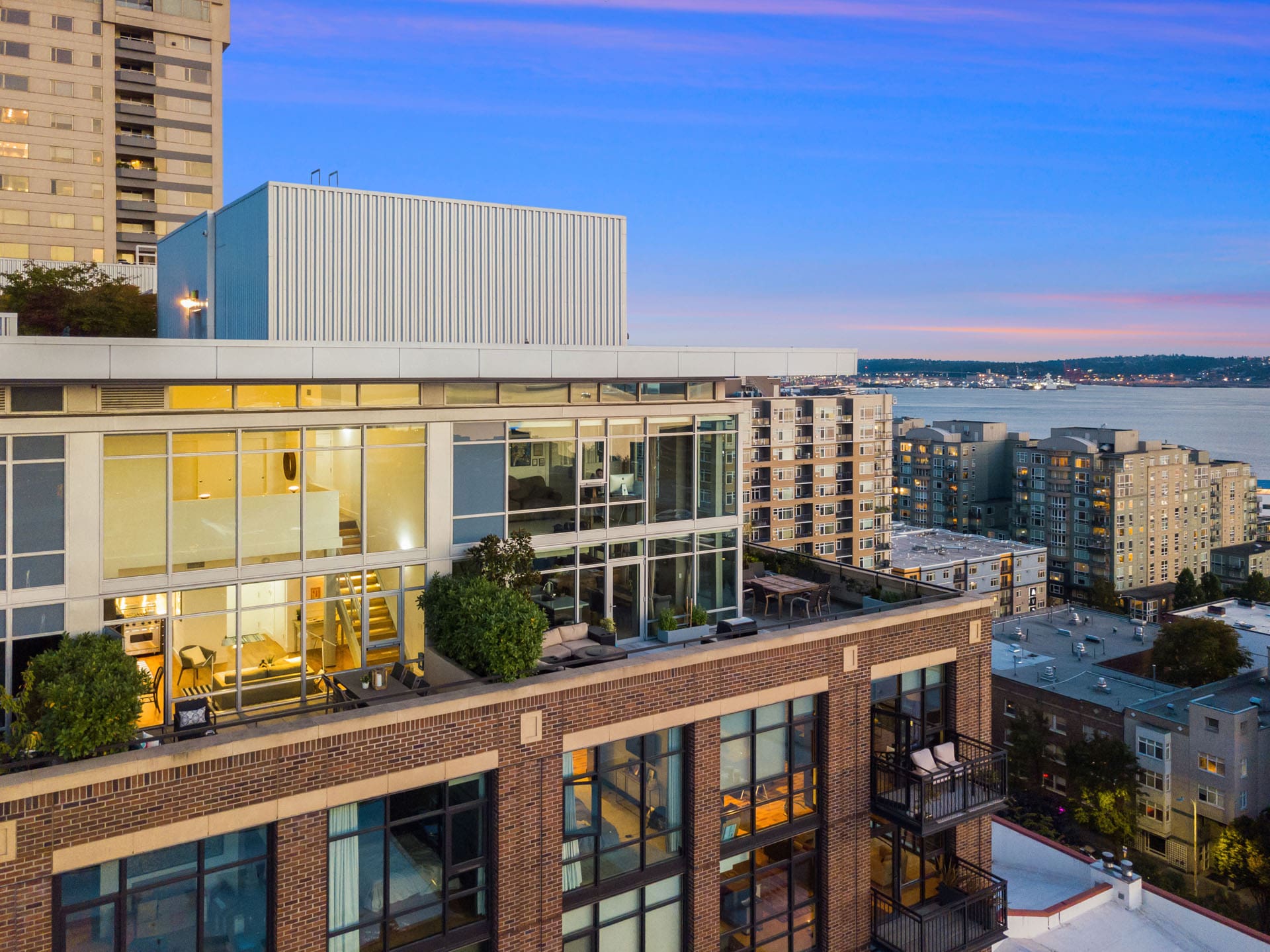 A modern apartment building with large glass windows stands prominently against the backdrop of a coastal cityscape. The sky is transitioning from sunset to twilight, casting a serene glow over the buildings and the distant water.