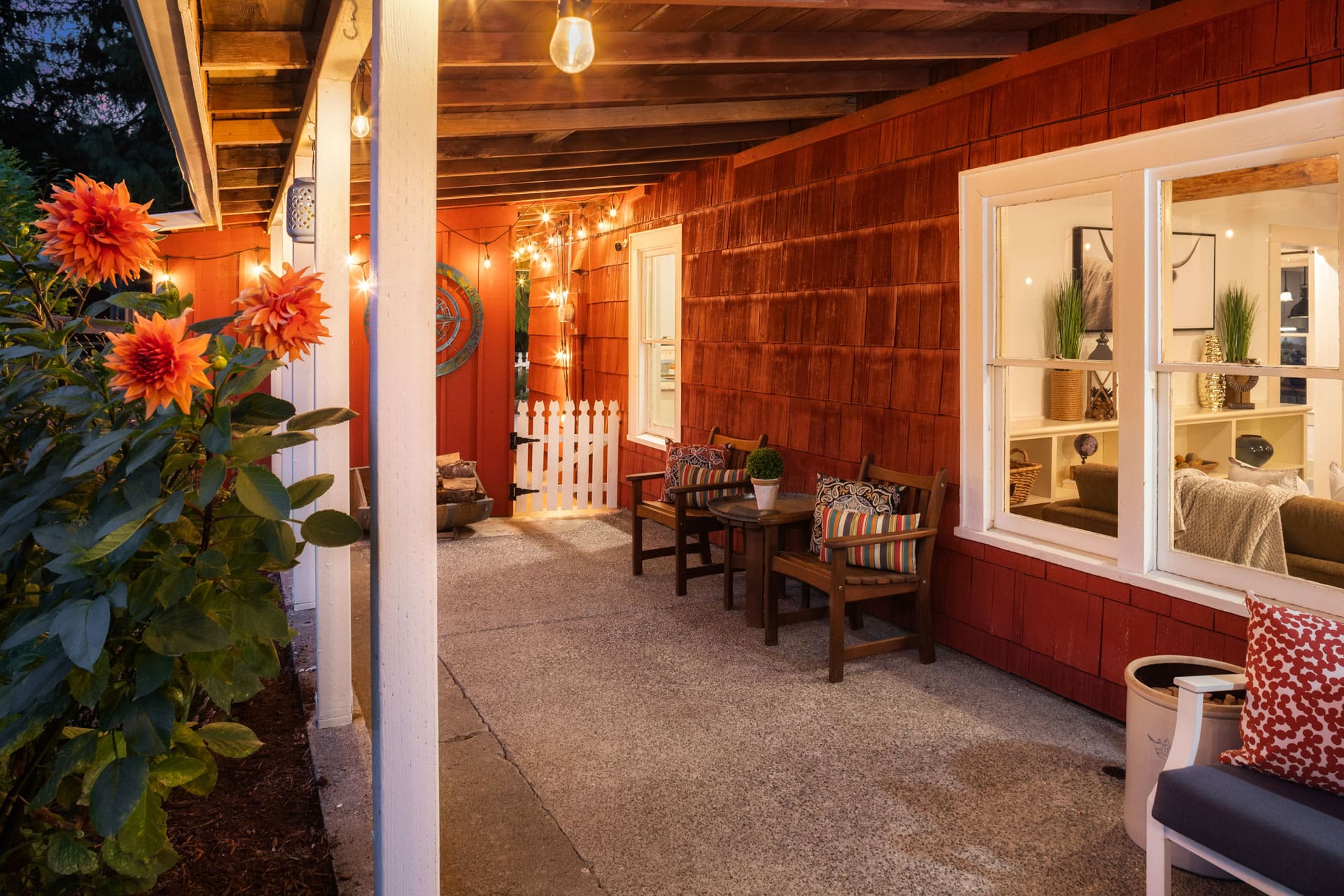 A cozy outdoor patio at dusk with warm lighting. The area features two wooden chairs with decorative cushions, a small table, potted plants, and string lights hanging from the roof. Bright orange dahlias are blooming to the left, near the entrance.