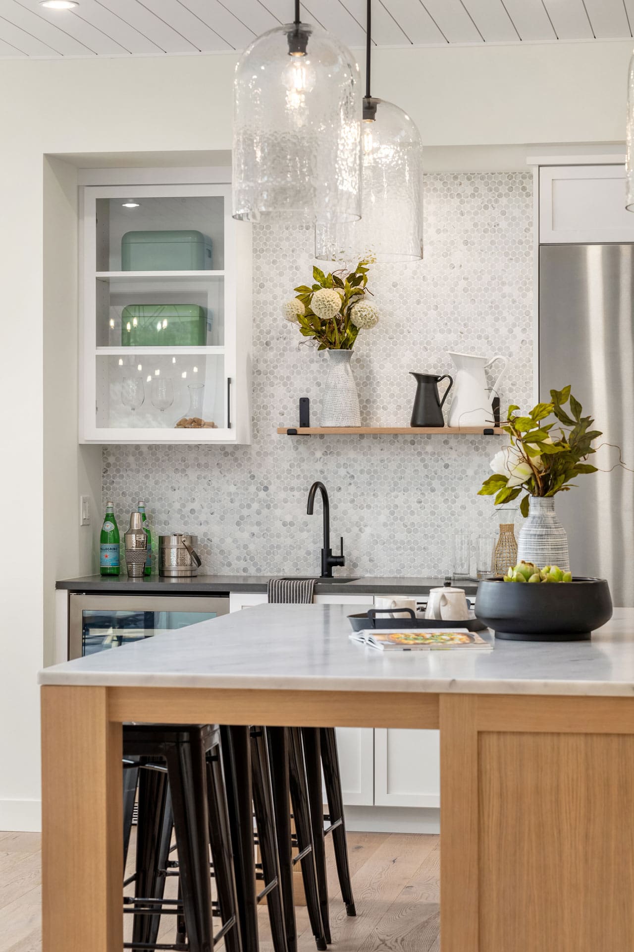 A modern kitchen features a light wood island with black stools underneath, a white marble countertop, and pendant lights above. The backsplash is a tiled mosaic, and the cabinets are white with glass doors. Greenery accents and kitchen essentials are neatly organized.