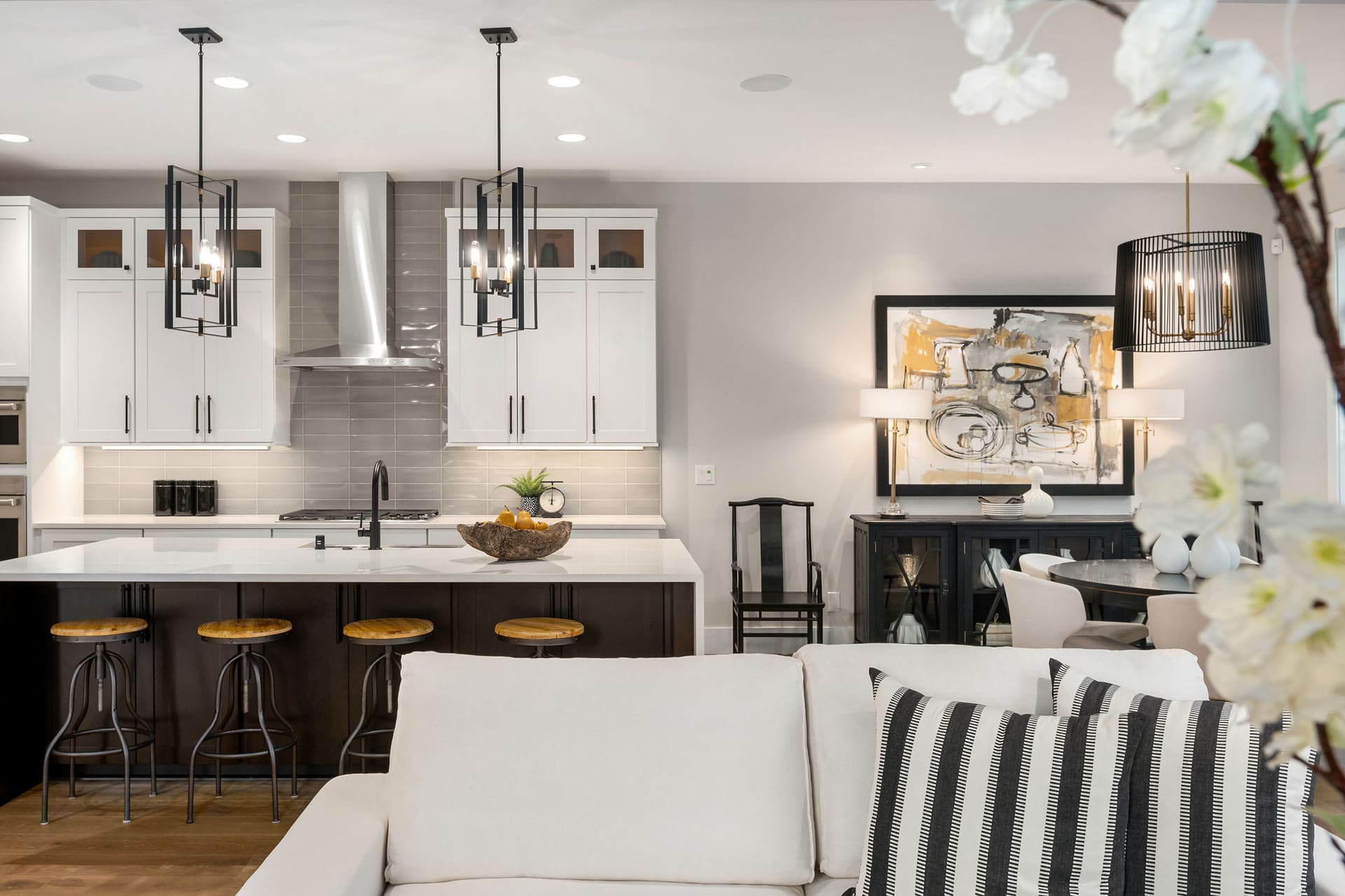 Modern kitchen and living area with sleek white cabinets, an island with wooden bar stools, pendant lighting, and stainless steel appliances. In the living area, there's a white sofa with striped pillows, abstract art on the wall, and contemporary decorative items.