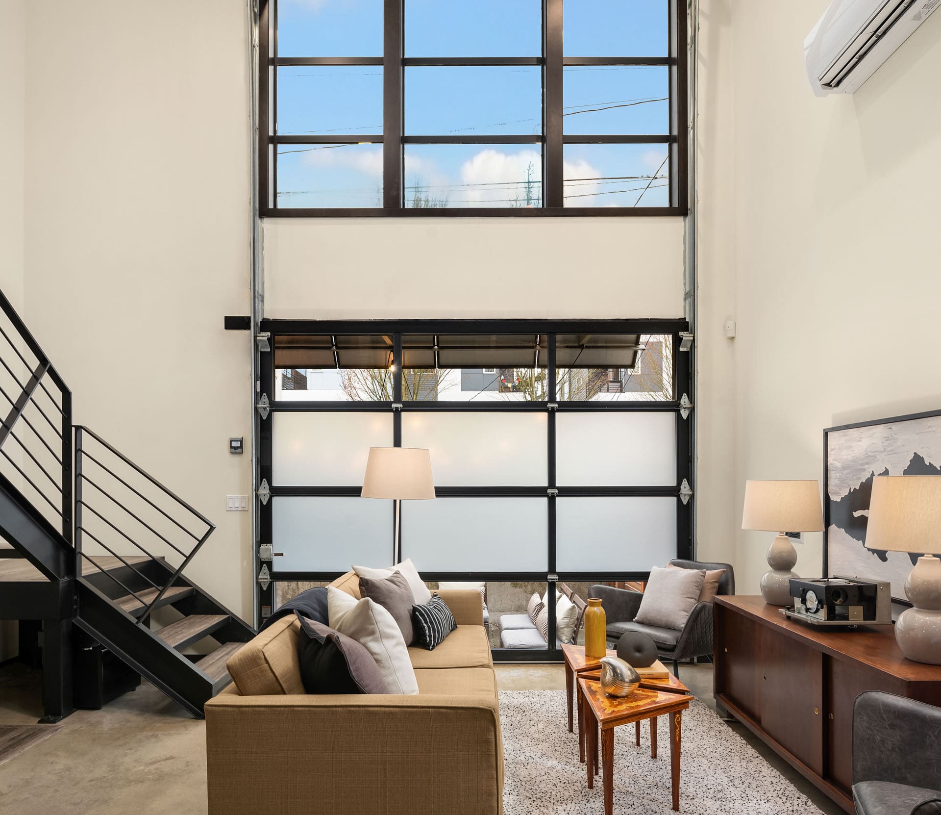 A modern living room with high ceilings and large windows. The space features a beige sofa, two coffee tables with decor items, a floor lamp, and an air conditioner mounted on the wall. A staircase with a black metal railing leads to an upper level.