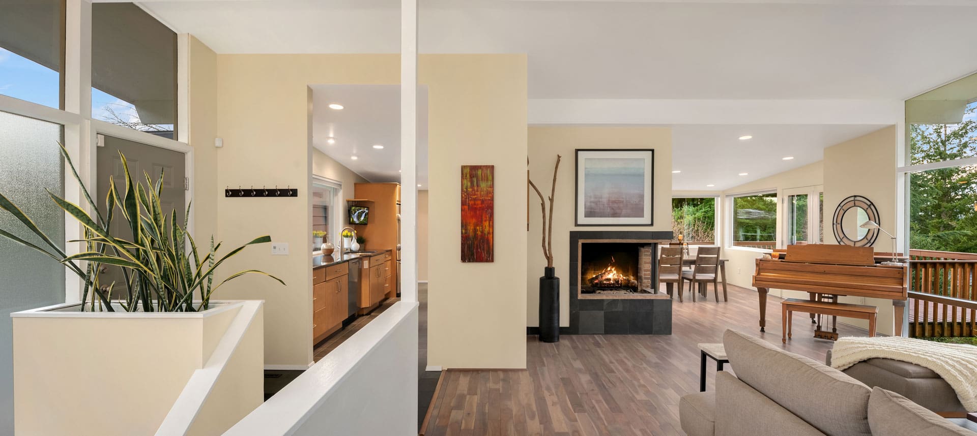 A modern open-concept living space features warm beige walls, recessed lighting, and hardwood floors. There is a kitchen with wooden cabinets, a dining area, and a fireplace adorned with a nature painting. A grand piano sits near large windows overlooking a forested area.