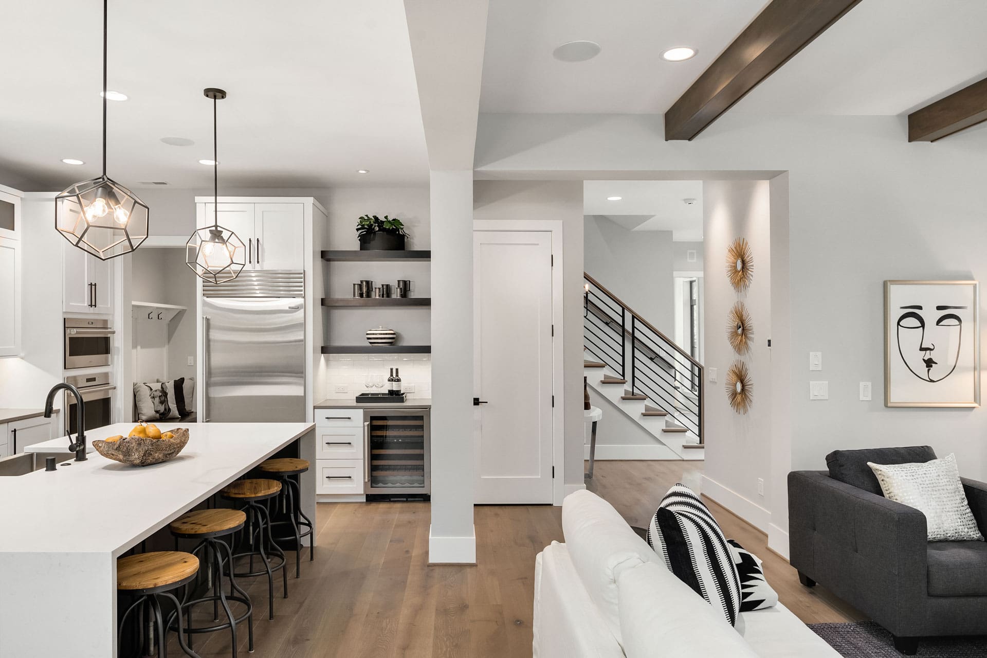 Modern open-plan living room and kitchen with white cabinetry, stainless steel appliances, and geometric pendant lights over the kitchen island. There are wooden stools at the island, a cozy seating area with a white sofa and dark gray chairs, and a staircase.