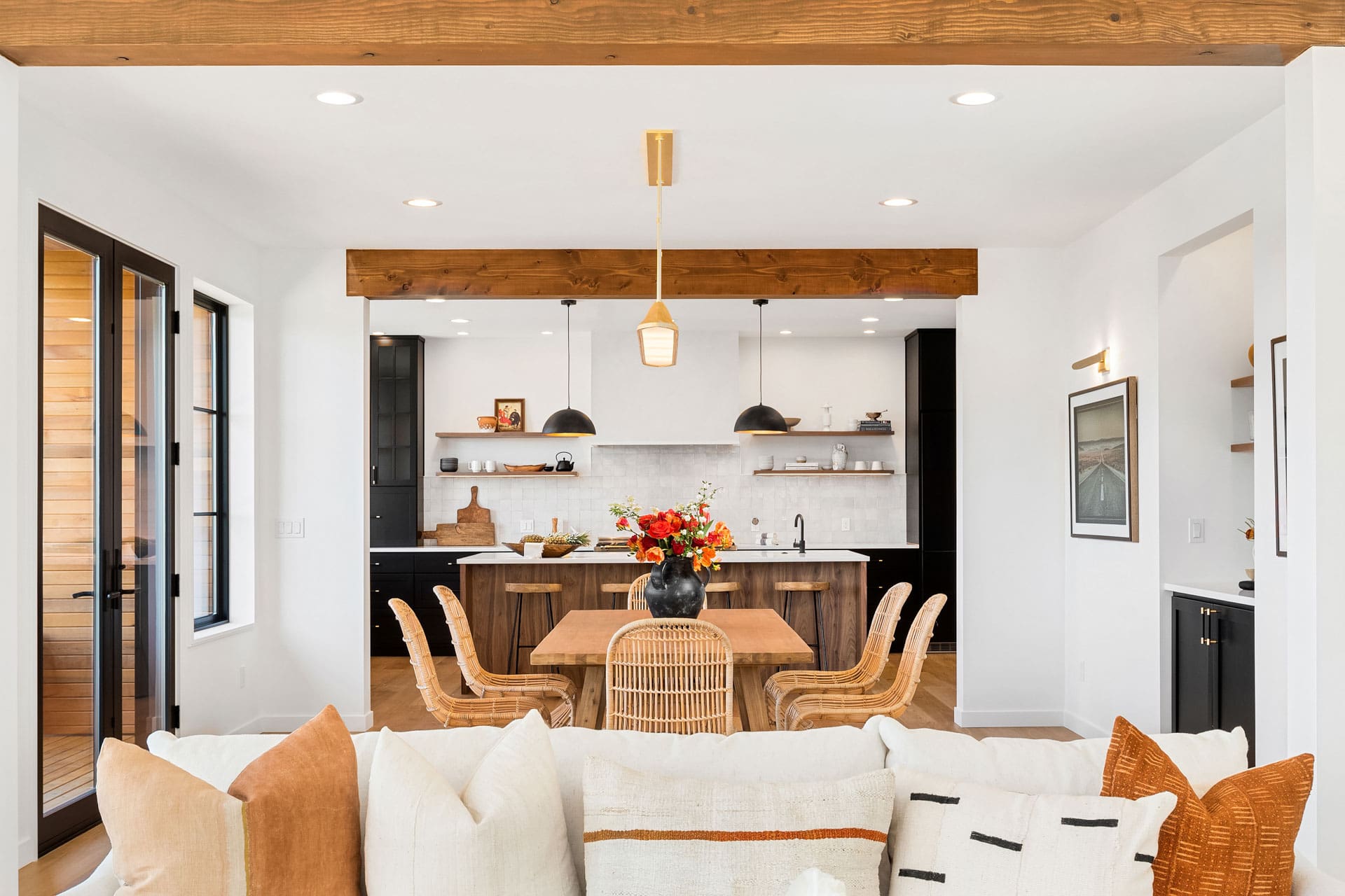 A modern and cozy open-concept living and dining area with a beige couch adorned with pillows in the foreground. The dining table is set with a floral arrangement, and the kitchen in the background features dark cabinets, white countertops, and wooden accents.