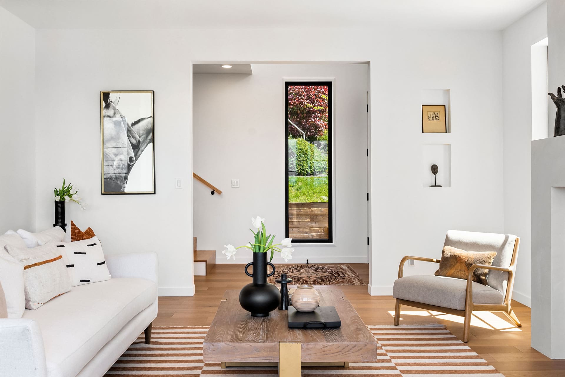 A modern living room with white walls and hardwood flooring features a white sofa with patterned cushions, a wooden coffee table with black vases, an armchair, decorative art, and a narrow window with a view of greenery outside. A staircase can be seen in the background.