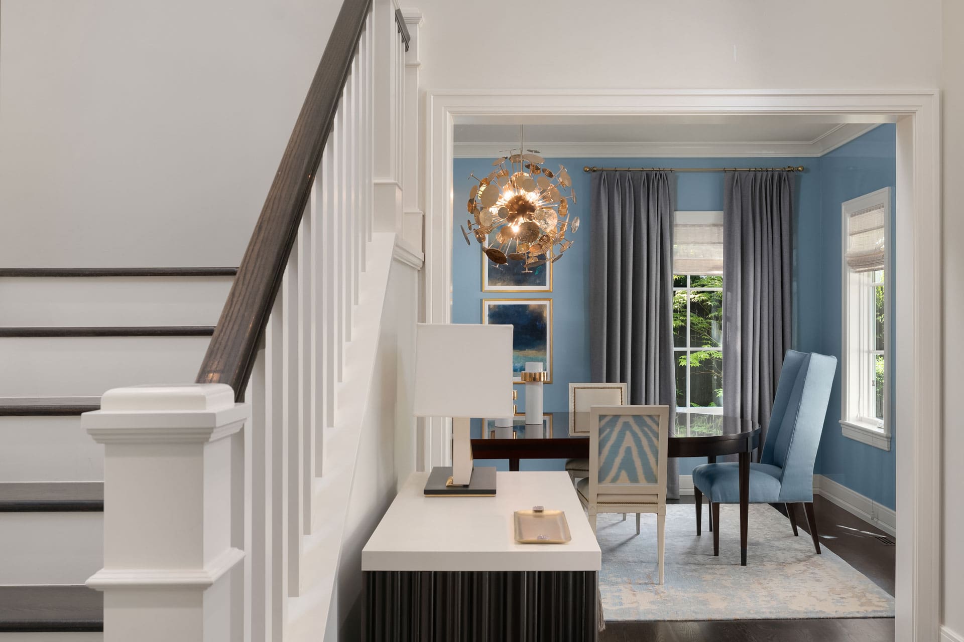 A modern interior features a staircase with white railings and dark-stained steps to the left. In the foreground, there's a stylish console table with a lamp. The adjacent dining room has blue walls, tall windows with gray curtains, and a contemporary chandelier.