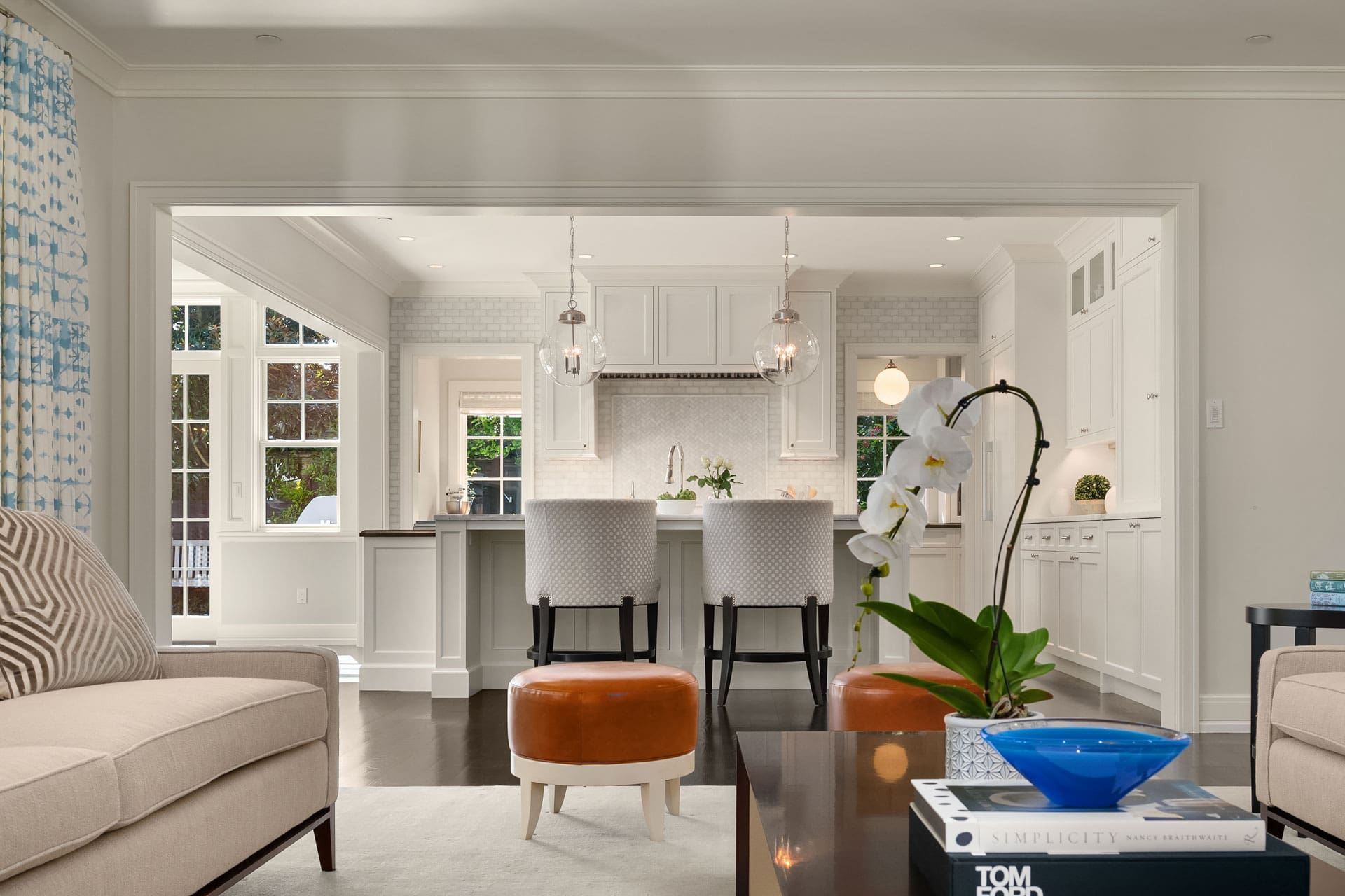 A modern living room seamlessly transitions into a bright, open kitchen. The living area has a beige sofa, two orange stools, and a coffee table with a white orchid and a blue bowl. The kitchen features white cabinetry, two bar stools at the counter, and pendant lights.