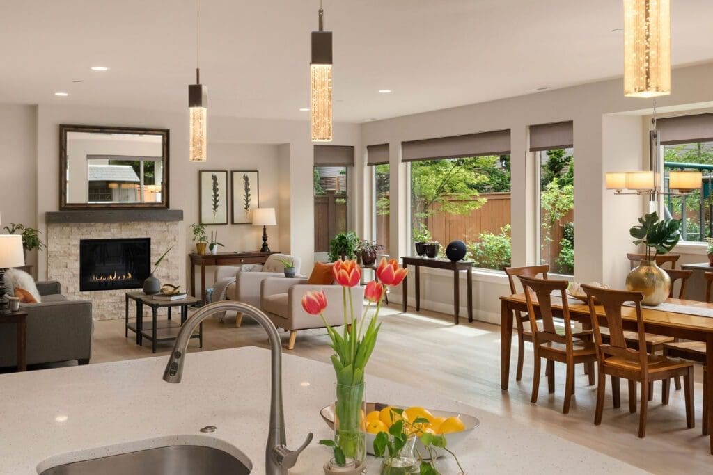 Real estate photography preparation with an open-plan living and dining area viewed from a modern kitchen. The room features large windows, contemporary furniture, and tasteful decor. Bright tulips in a vase sit on the kitchen counter, while the living area includes a fireplace and a cozy seating arrangement.