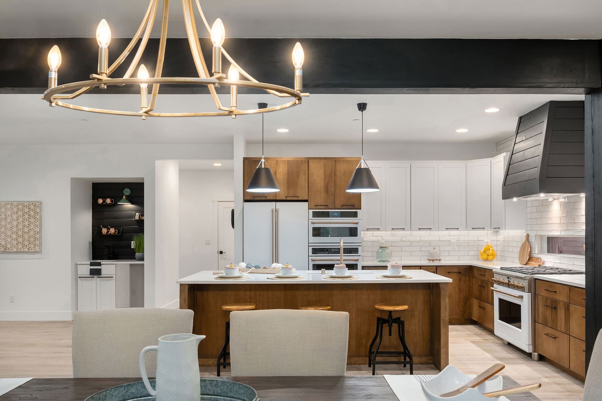 A modern kitchen and dining area with a large island. The kitchen features wooden cabinets, white countertops, stainless steel appliances, a dual oven, and a black range hood. The island has three stools and pendant lights above. A gold chandelier hangs above the dining table set with beige chairs.