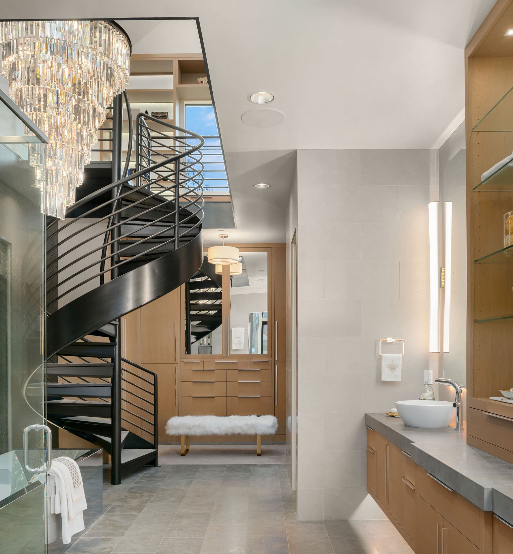 A modern, elegant bathroom featuring a sleek black spiral staircase leading to an upper level. The space includes wooden cabinetry, a large mirror, glass shelving, and a contemporary chandelier. Natural light fills the room, enhancing its sophisticated design.