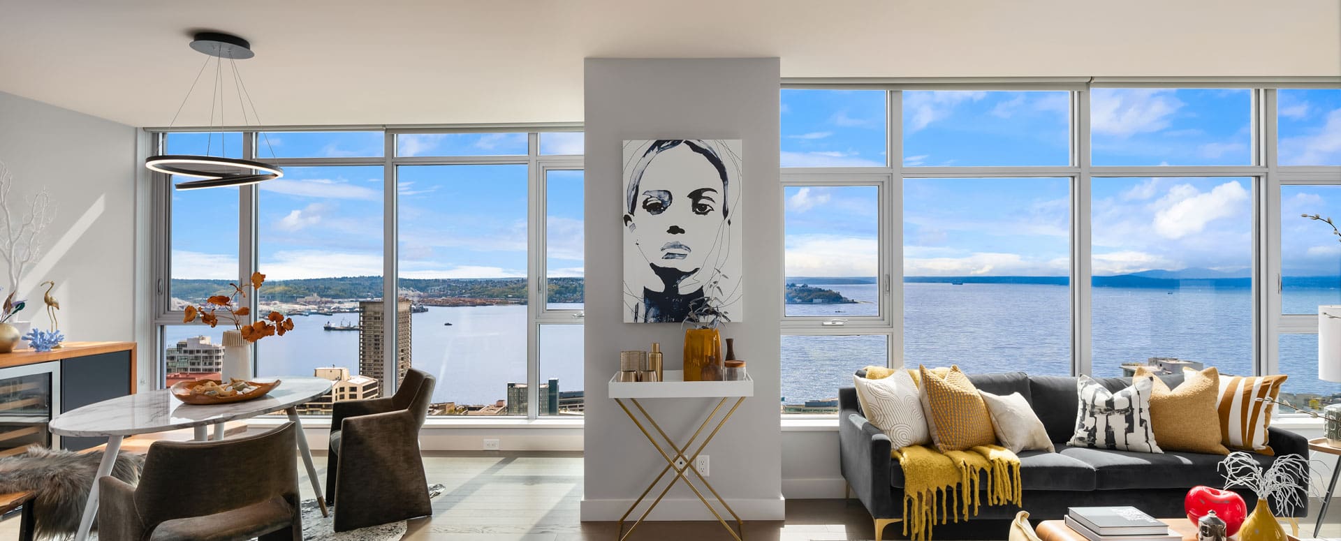 A modern, spacious living room with large windows offering a panoramic view of a serene body of water and distant islands. The room includes a circular dining table, a gray sofa with yellow accents, a wall-mounted shelf with a monochrome portrait, and contemporary decor.