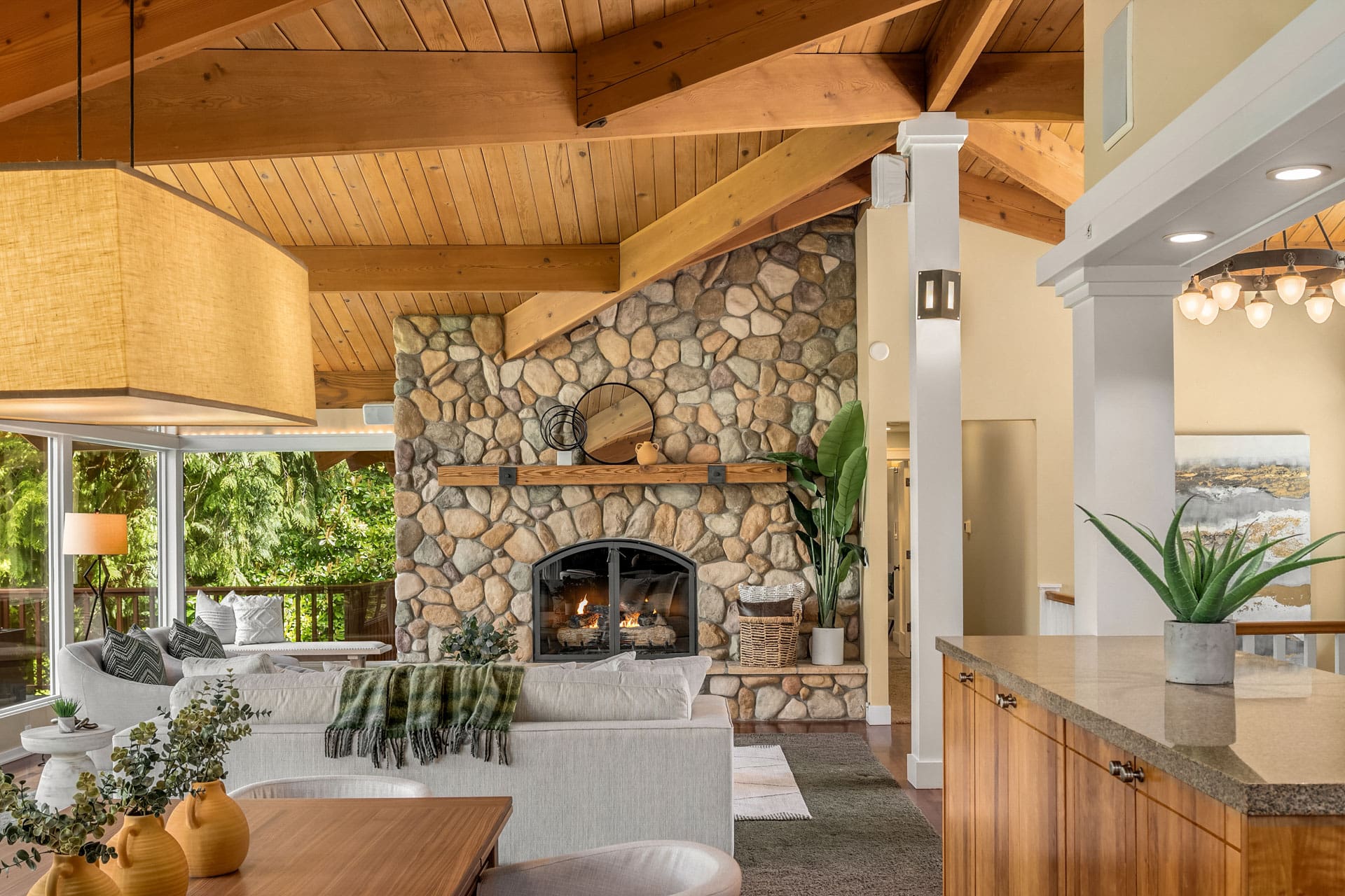 A cozy living room with a wooden vaulted ceiling and a stone fireplace. The room features a mix of contemporary and rustic decor, including a beige sofa, wooden dining table, and indoor plants. Large windows bring in natural light and offer a view of lush greenery outside.