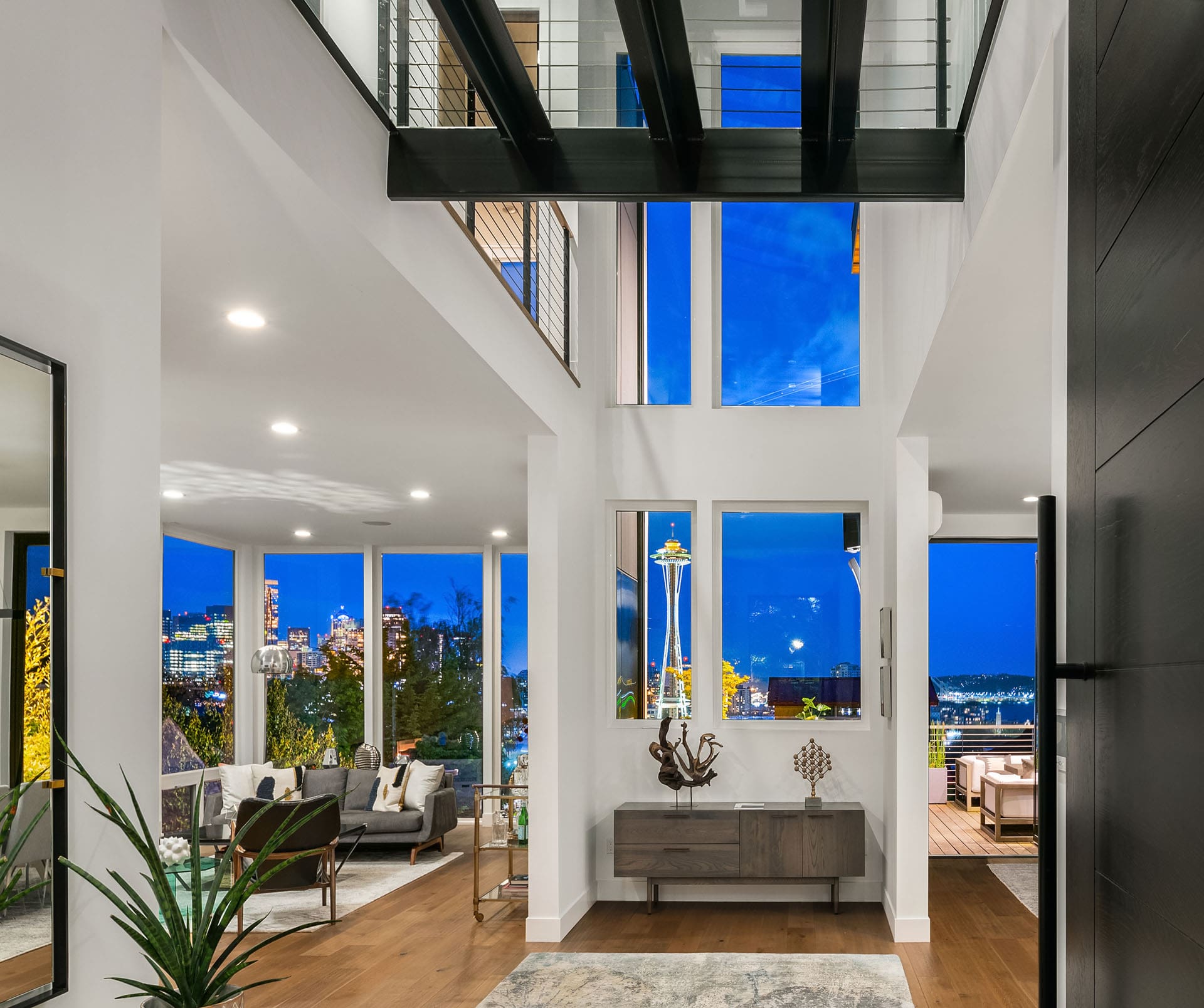 A modern, open-concept living area illuminated by bright recessed lighting. Large windows provide a stunning view of the city skyline at night. Stylish furnishings, a wooden console, and indoor plants enhance the contemporary ambiance.