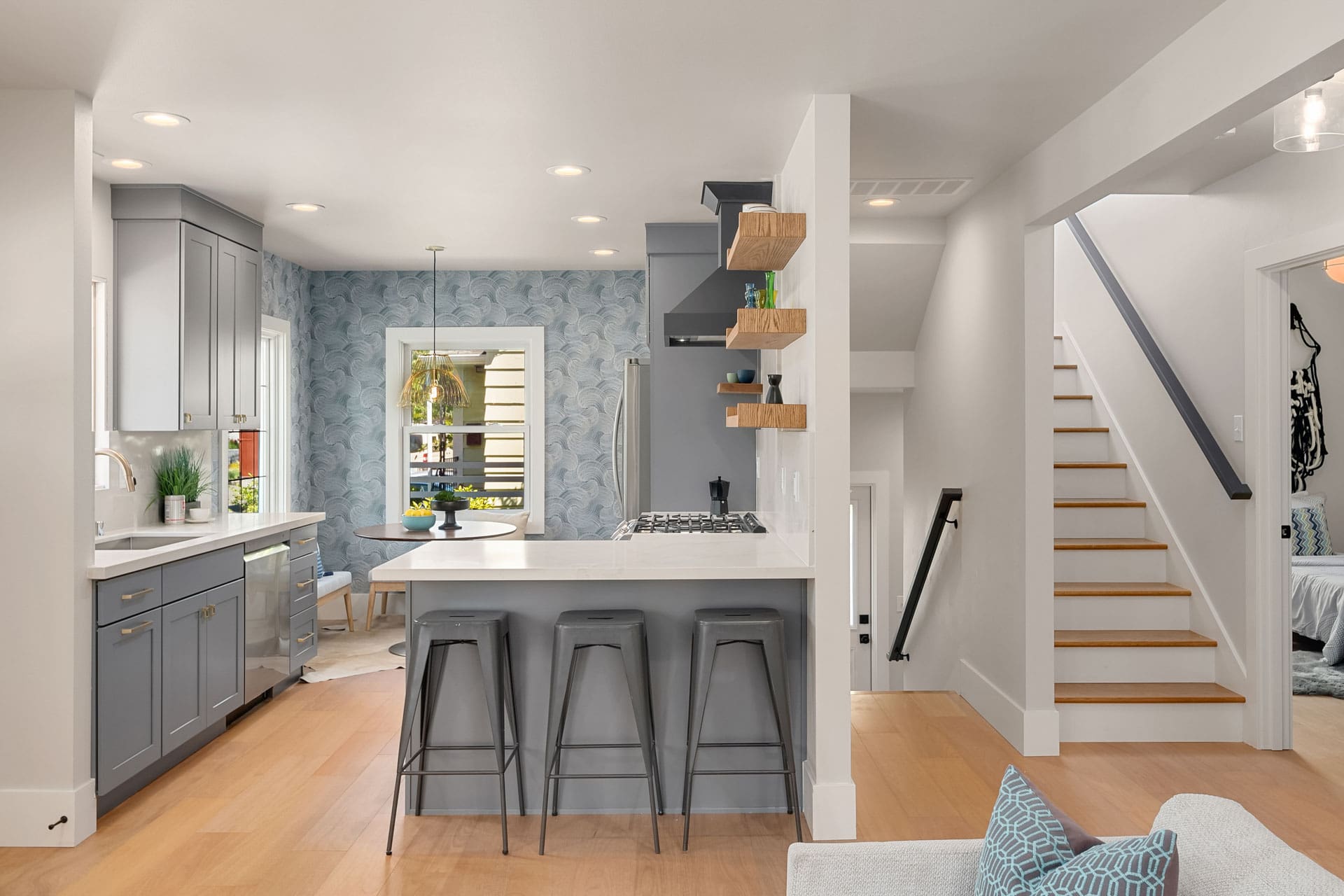 Modern kitchen with a white island counter and three gray barstools. The kitchen has blue cabinets, wood floating shelves, and a small dining area with a patterned wallpaper accent wall. A staircase with light wood steps and a dark railing leads to the upper floor.