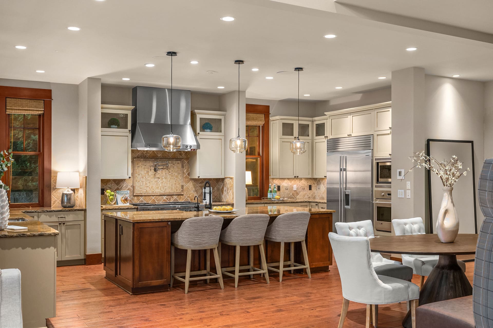 Modern kitchen and dining area featuring a central island with four cushioned bar stools, stainless steel appliances, and a tiled backsplash. Pendant lights hang above the island. The dining table is set with six chairs and decorative elements. Warm wood flooring throughout.