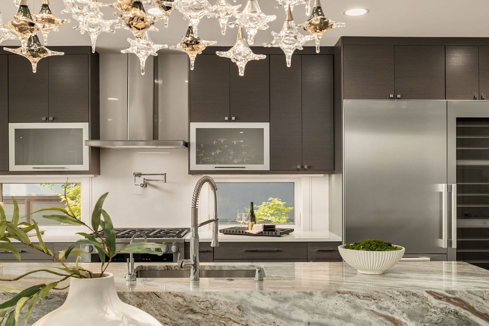 Modern kitchen with dark wood cabinets and stainless steel appliances. A large island features a marble countertop with a sink and faucet. A plant and a bowl with greenery are on the countertop. A chandelier with star-shaped ornaments hangs above.