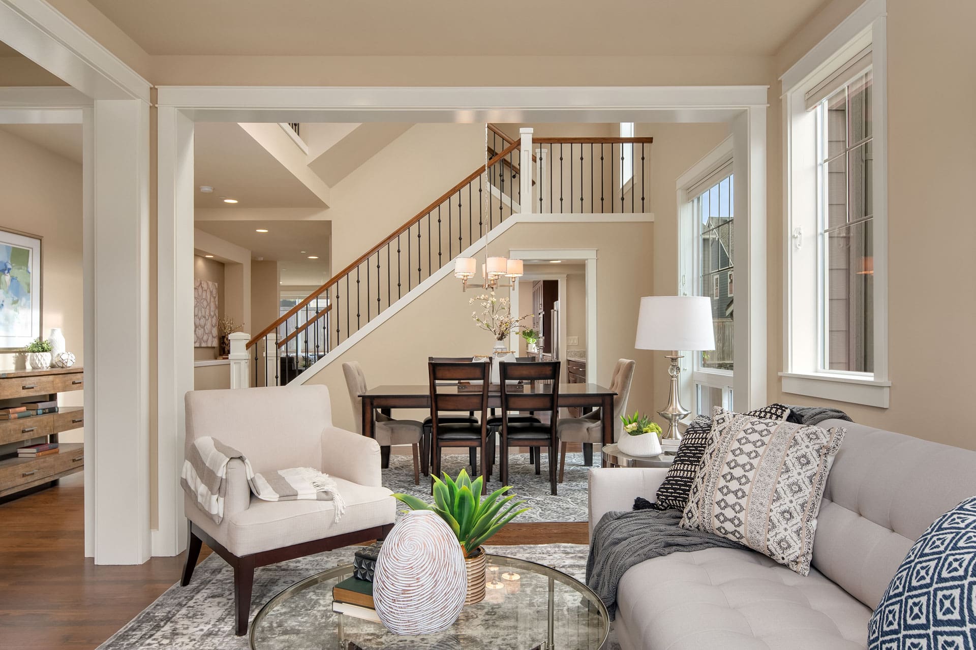 A modern living room with neutral tones, featuring a gray sofa with patterned pillows, a beige armchair, and a glass coffee table with decorative items. A dining area with a dark wood table and chairs is in the background, near a staircase with wooden steps.