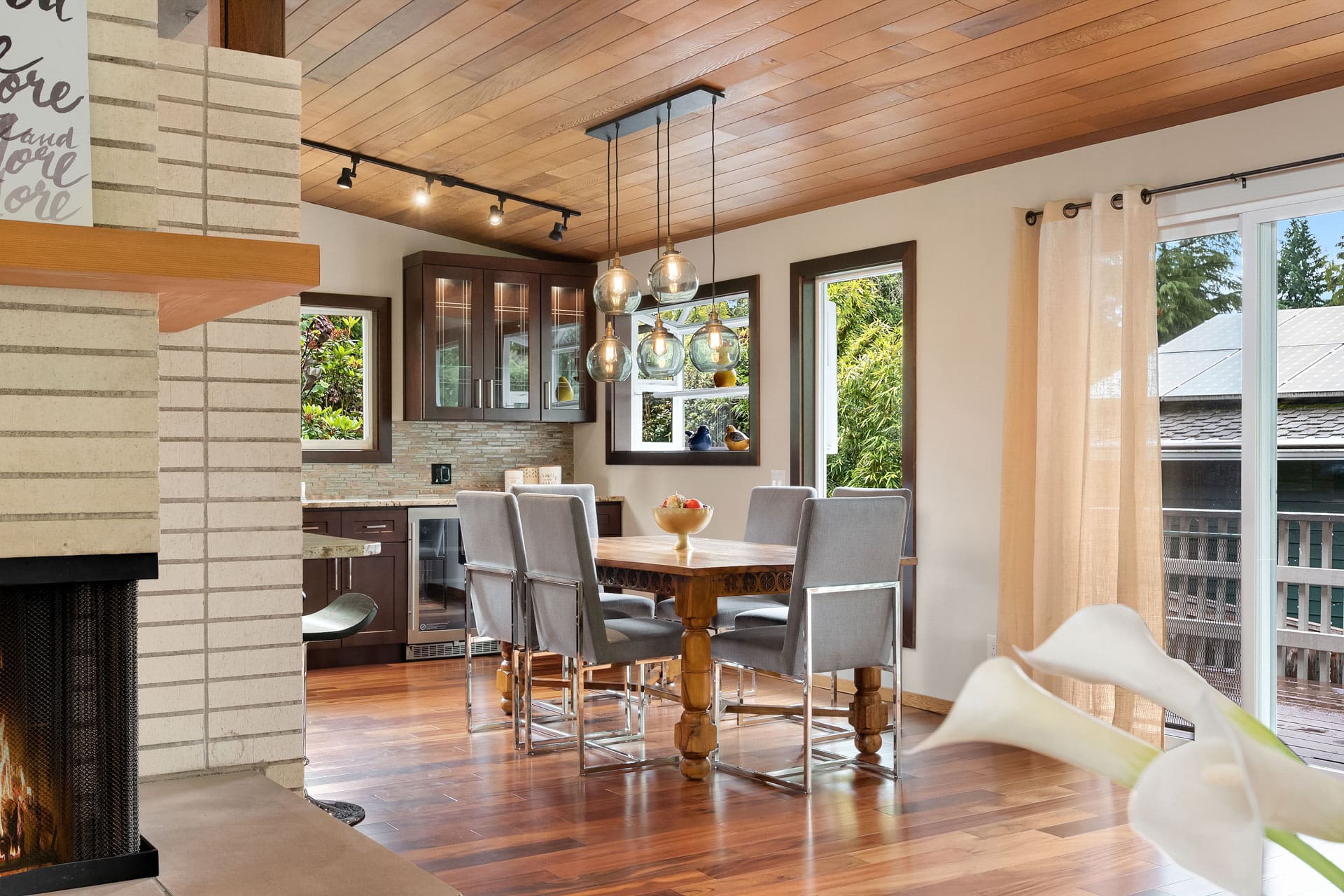 A cozy dining area with a wooden table surrounded by grey cushioned chairs, set in an open-plan room with wooden flooring and ceiling. Large windows and sliding glass doors let in natural light, offering a view of a deck and greenery outside. A modern kitchen is visible in the background.