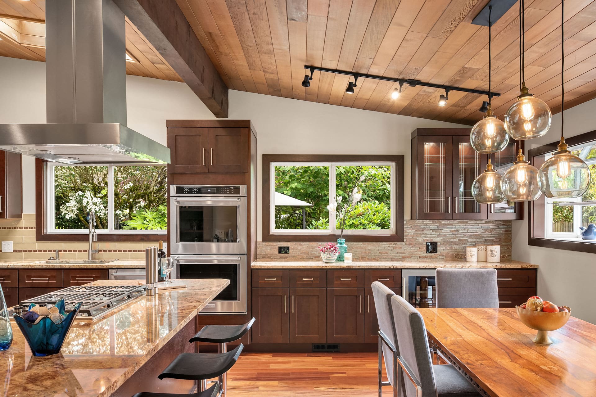 A modern kitchen with wooden cabinetry, stainless steel appliances, and granite countertops. The kitchen island has bar stools and a built-in stove. Pendant lights hang above a wooden dining table. Large windows offer views of greenery outside.